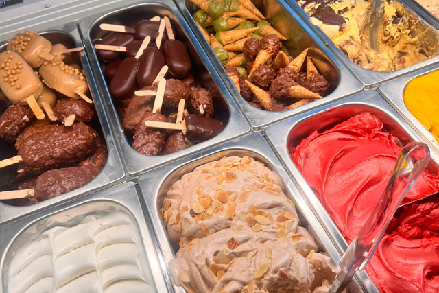 Gelato assortito in vaschette di metallo, tra cui gusti al cioccolato, vaniglia, frutta e rosso acceso, con paletta e spatola.