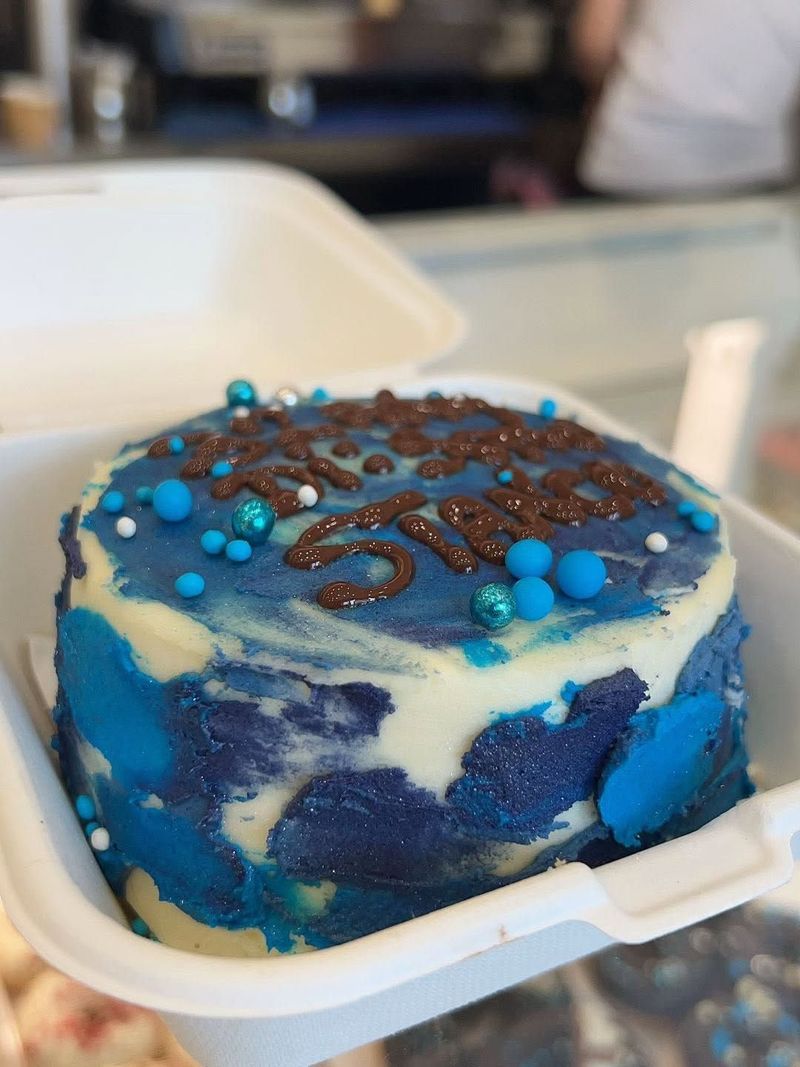 Torta con glassa blu, glassa al cioccolato e zuccherini blu, servita in un contenitore bianco da asporto.