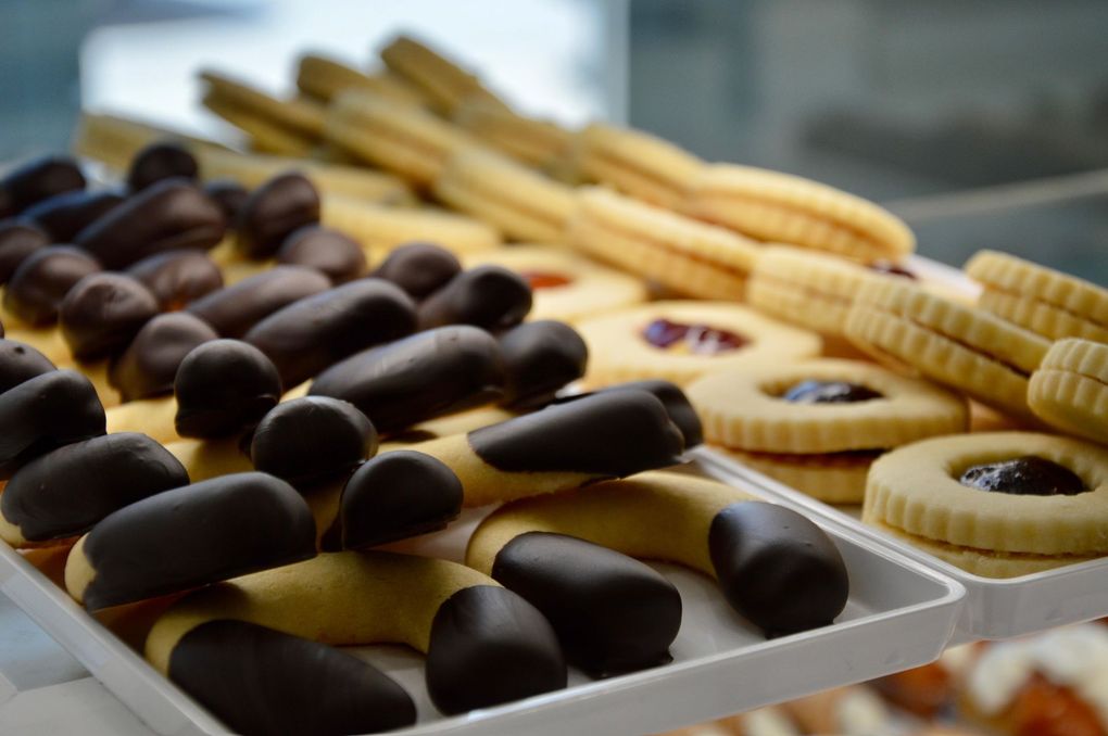 Assortimento di biscotti e frollini ricoperti di cioccolato, disposti su vassoi in una vetrina di una pasticceria.