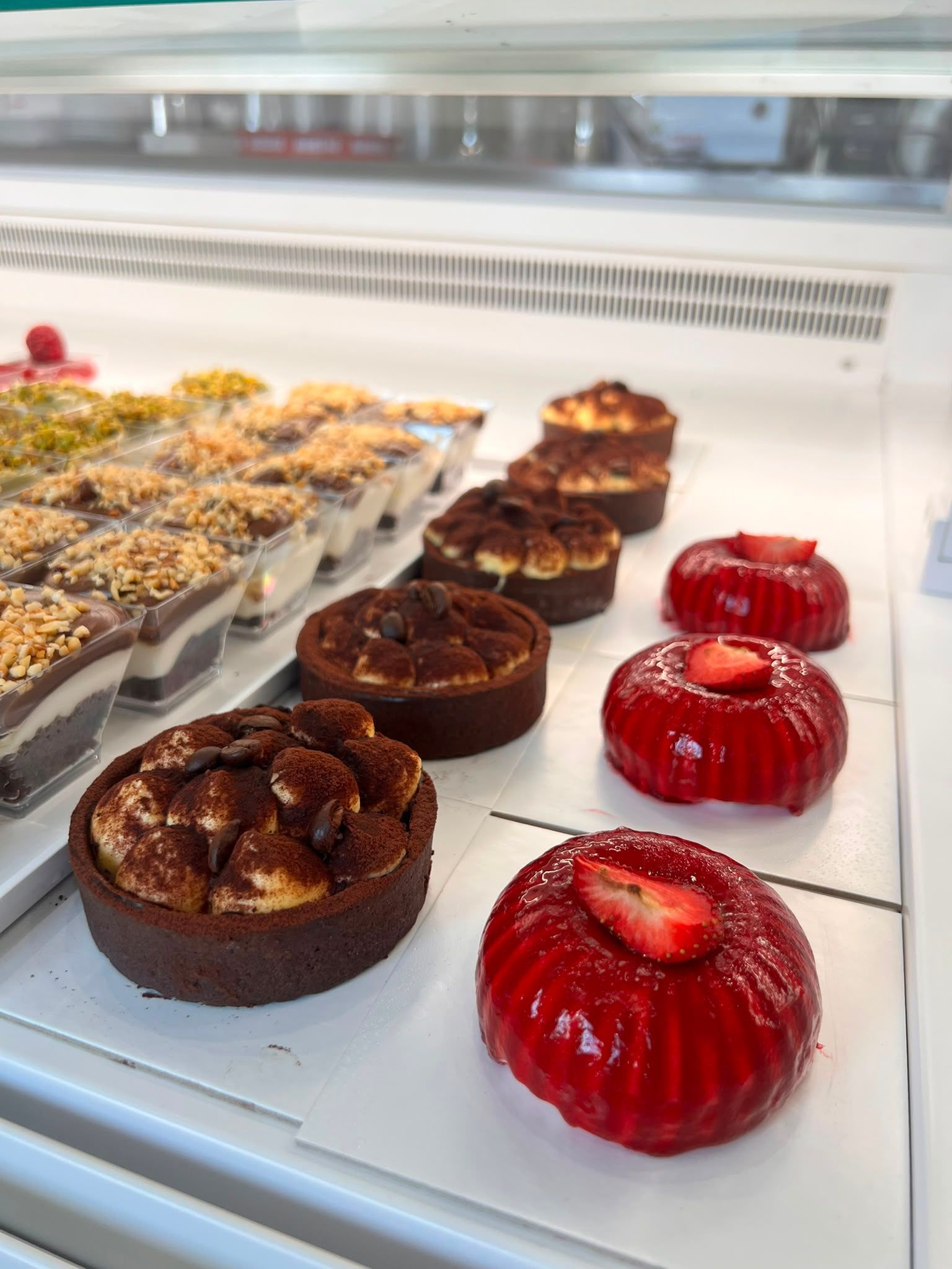 Assortimento di pasticcini in una vetrina da pasticceria, tra cui tortine glassate di rosso e crostatine al cioccolato.