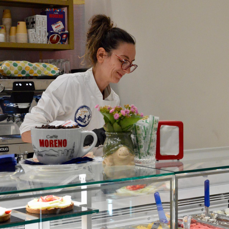 Un barista in giacca bianca sorride dietro un bancone di vetro ornato da fiori, tazze e attrezzatura per il caffè.