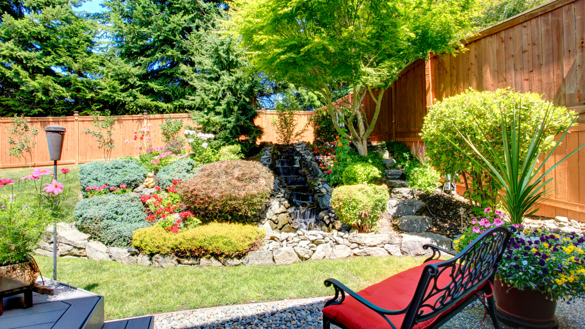 Backyard garden with a waterfall, a wooden fence, and a decorative wrought iron bench.