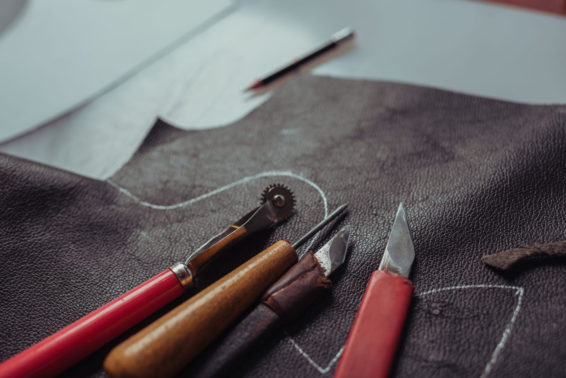 Attrezzi per la lavorazione del cuoio, tra cui una rotella da ricalco, un punteruolo e un taglierino, sono appoggiati su un pezzo di cuoio scuro con disegni incisi.