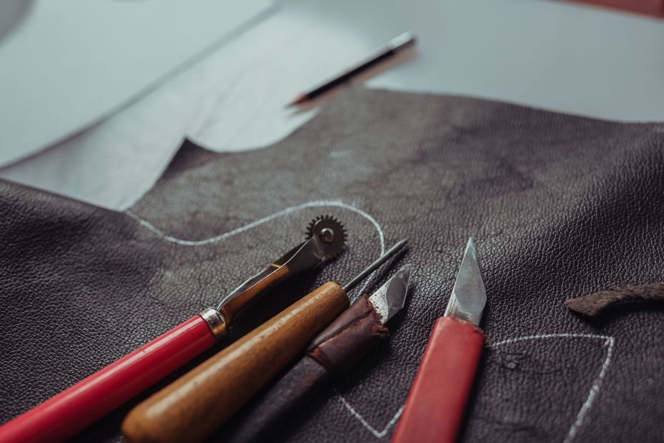Attrezzi per la lavorazione del cuoio, tra cui una rotella da ricalco, un punteruolo e un taglierino, sono appoggiati su un pezzo di cuoio scuro con disegni incisi.