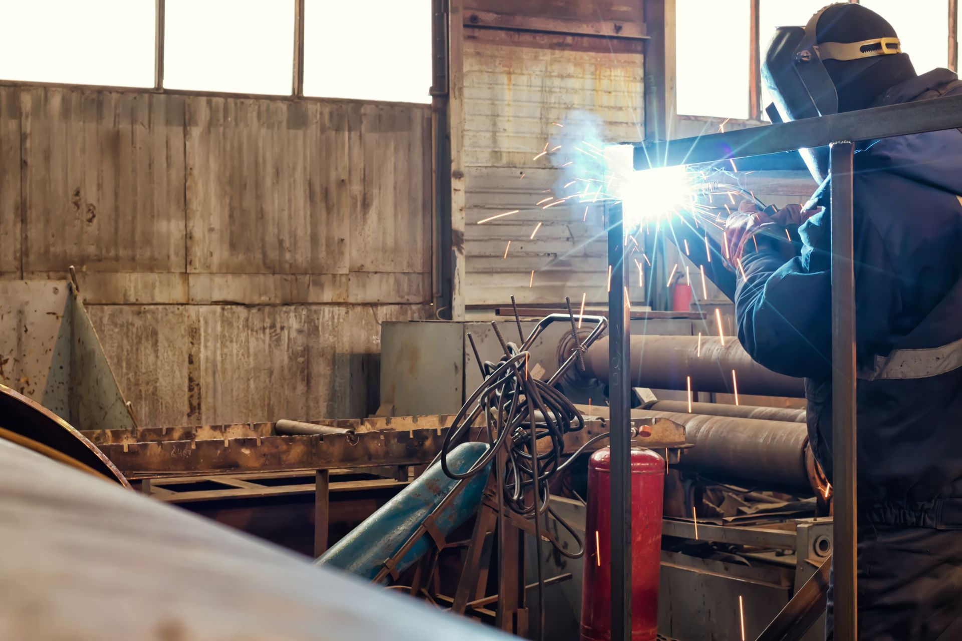 Welder in a workshop sparks bright light while working on metal machinery amid pipes and tools.