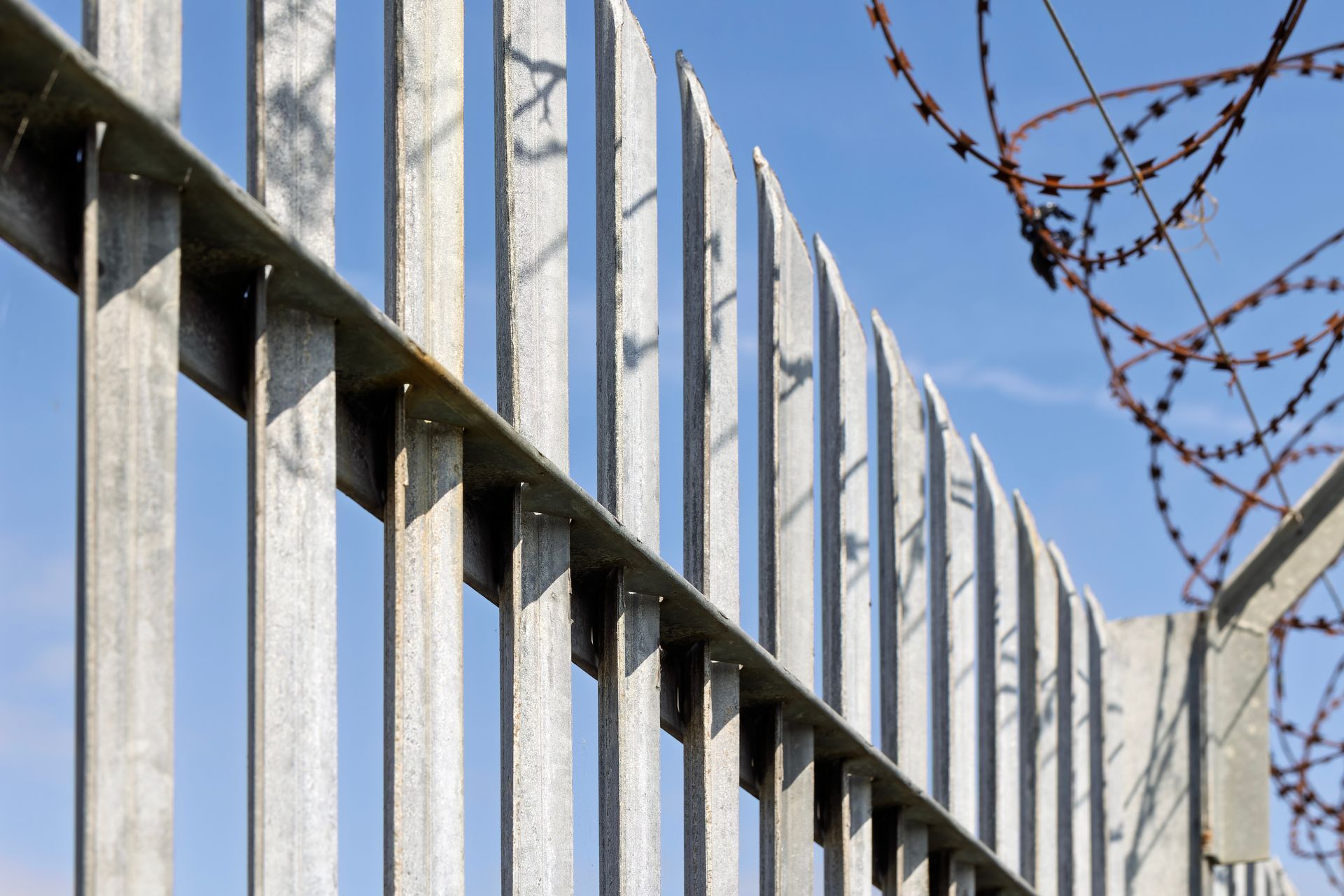 Barbed wire atop a tall metal fence against a blue sky