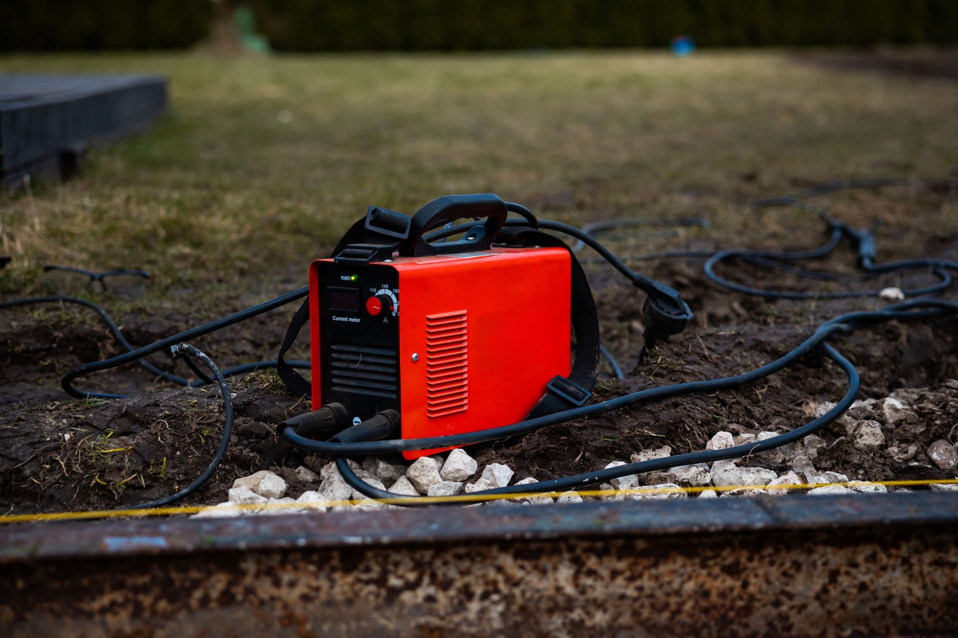 Red portable welding machine on wet ground with cables and white rocks nearby outdoors