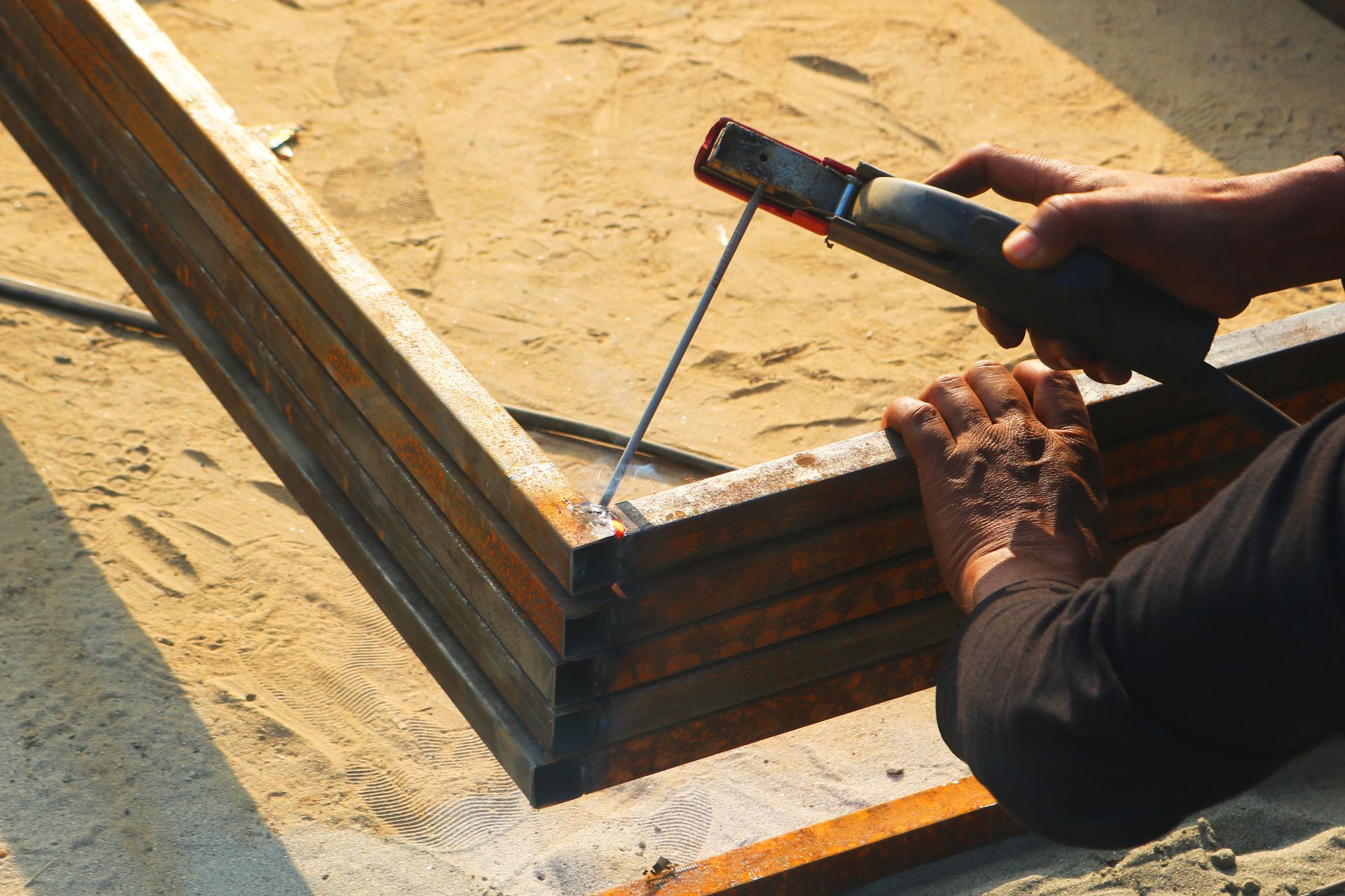 Worker welding a rusted metal frame in bright sunlight, with sparks and a handheld torch.