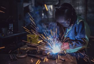 Welder in blue protective gear welding metal, with bright sparks in a dark workshop.