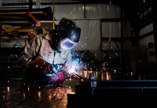 Welder in protective gear sparks metal in a dim workshop, bright blue arc lighting the scene