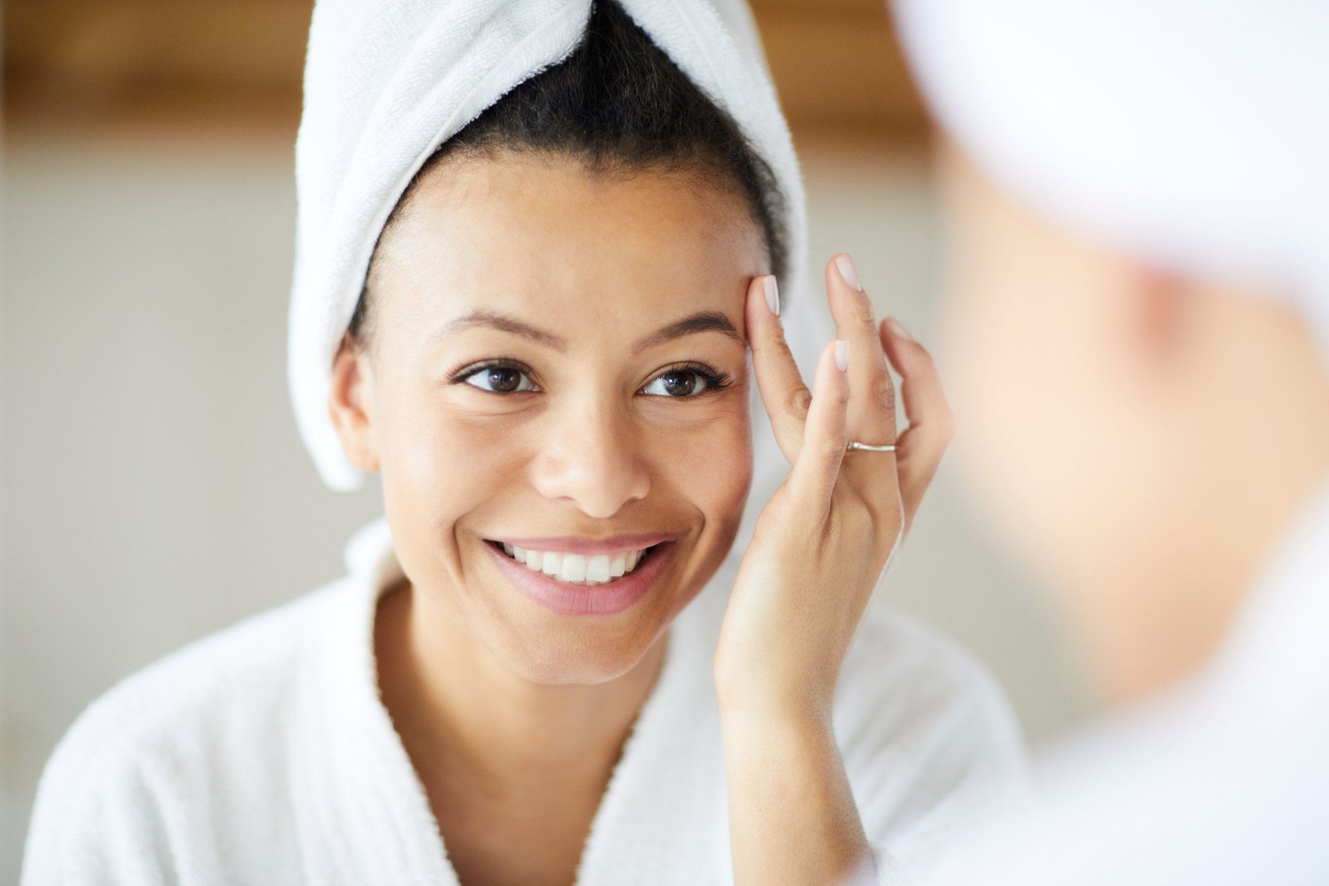 A woman with a towel wrapped around her head is looking at her face in the mirror.