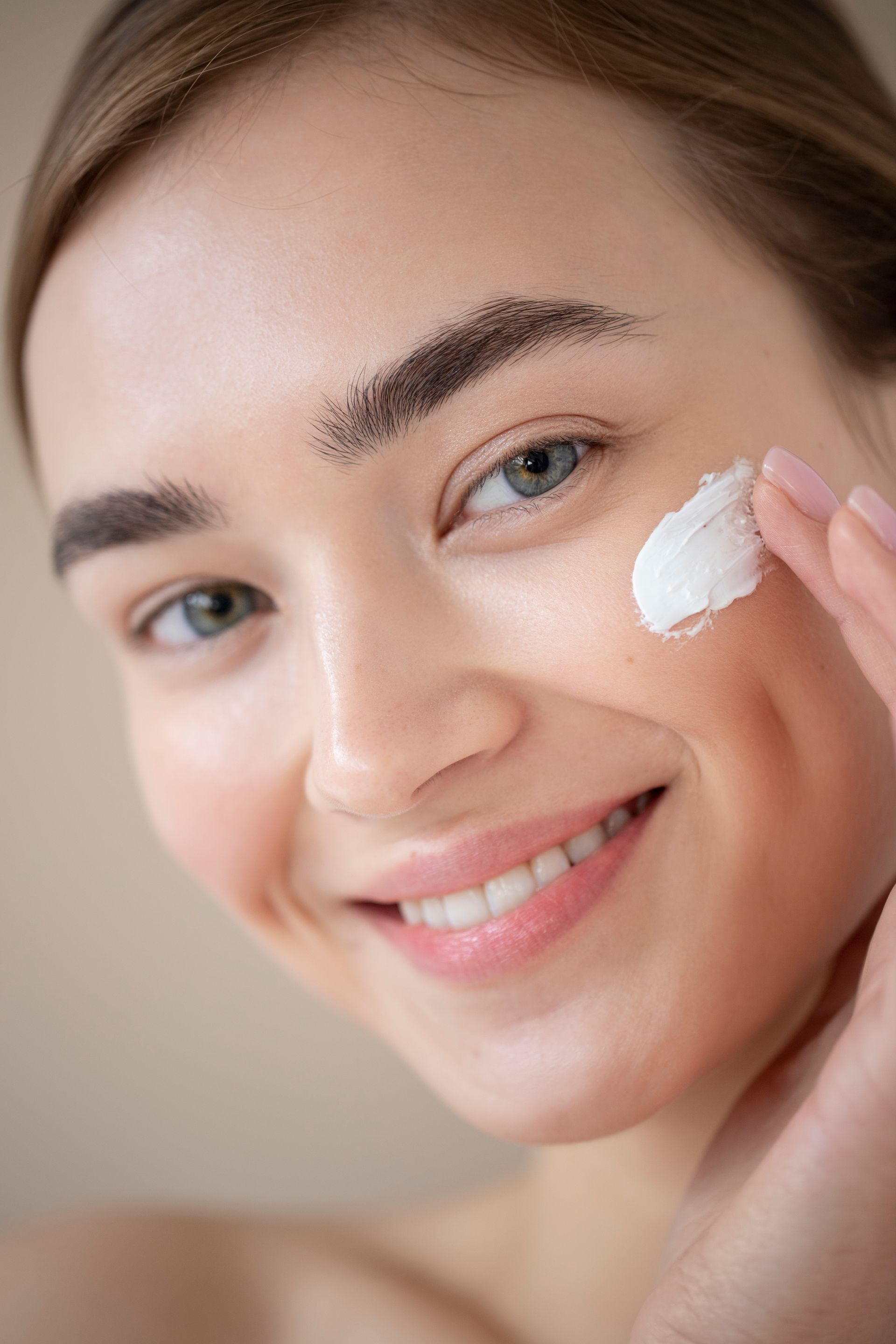 A woman is smiling while applying cream to her face.