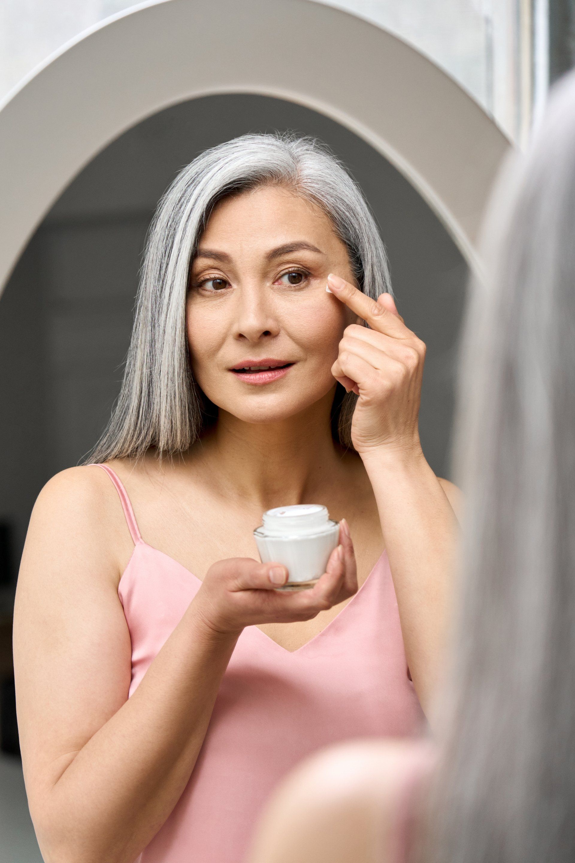 A woman is applying cream to her face in front of a mirror.