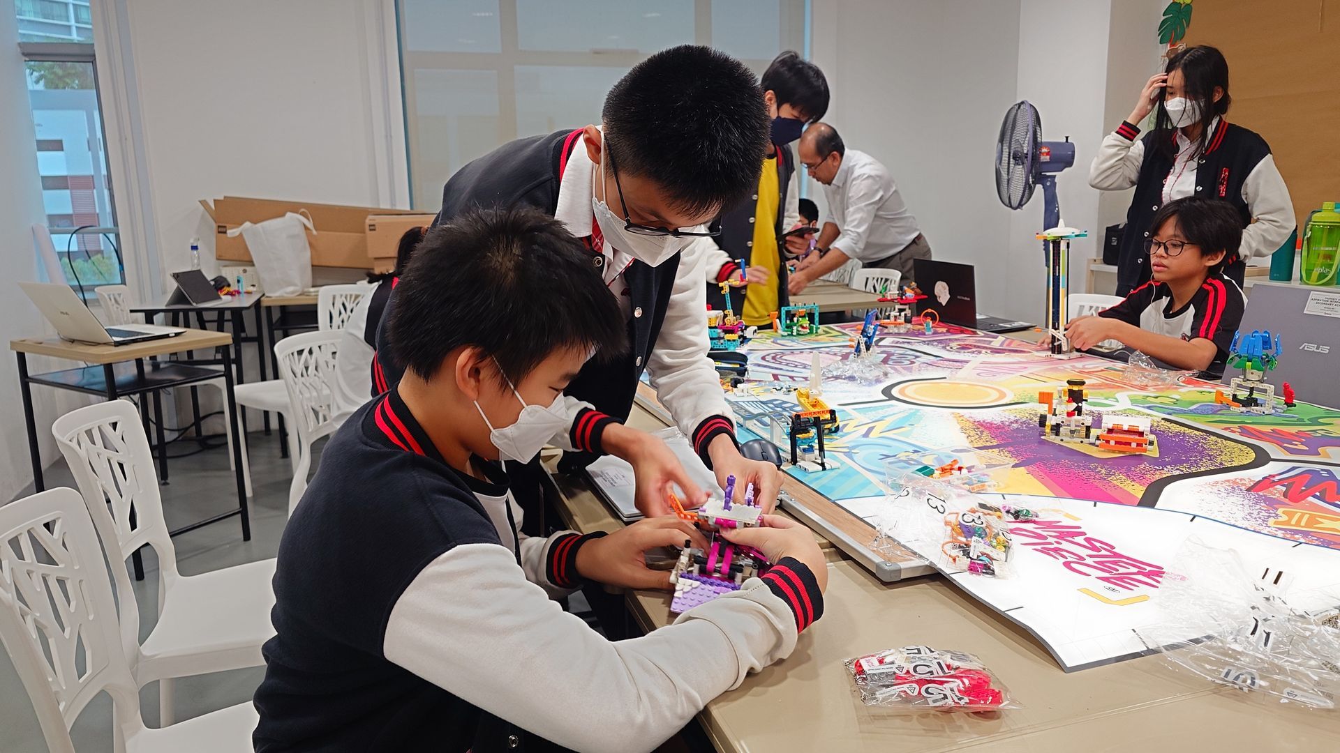 Students wearing masks collaborate on a colorful, sprawling craft project at a table in a brightly lit room.