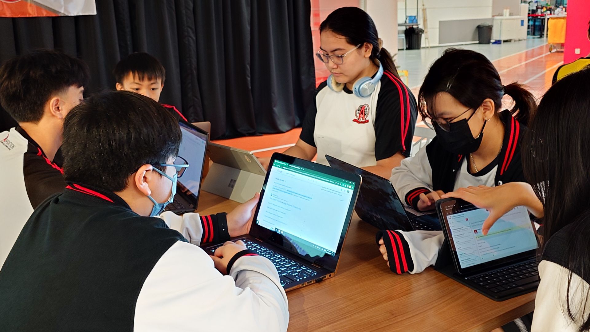 Students in matching school uniforms sit at a table working together on laptops and tablets in a brightly lit indoor space.