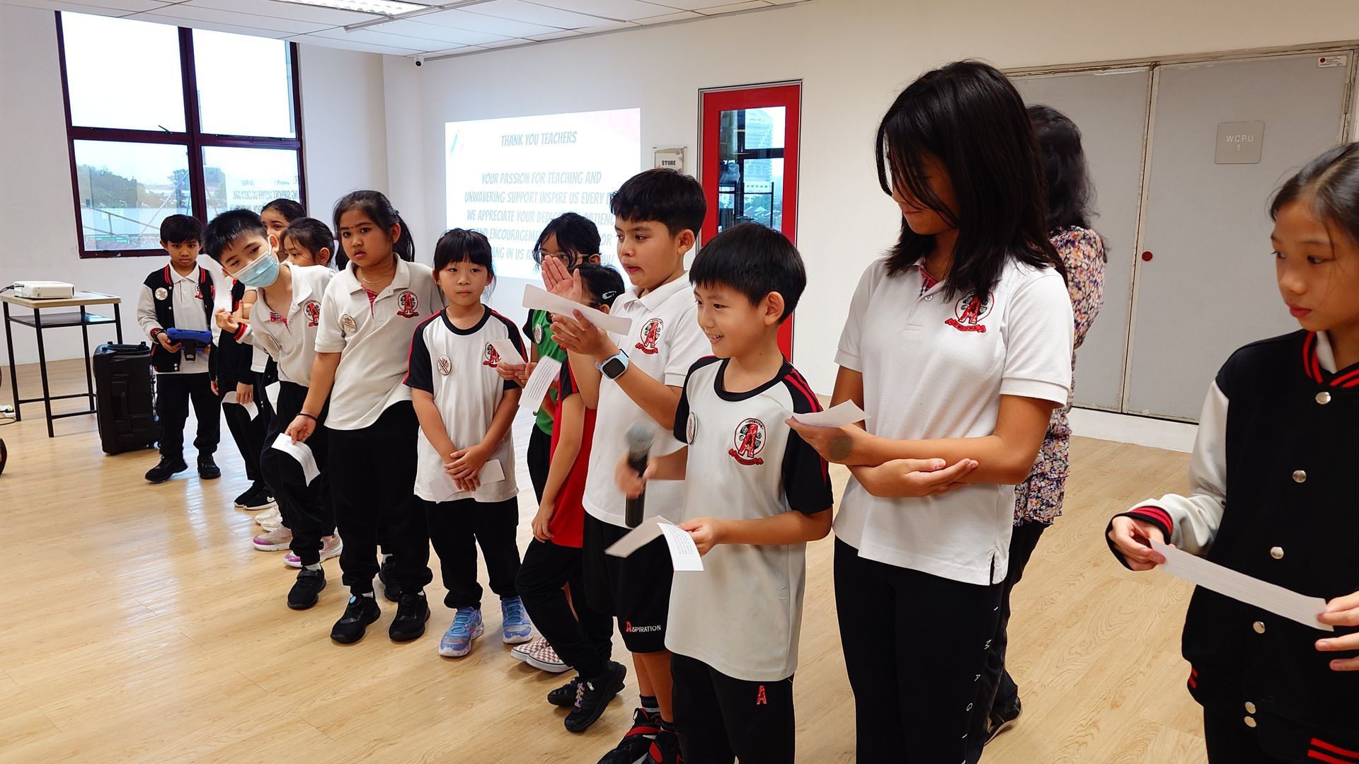 A group of students in uniforms standing in a line in a classroom, with some holding papers, during an indoor activity.
