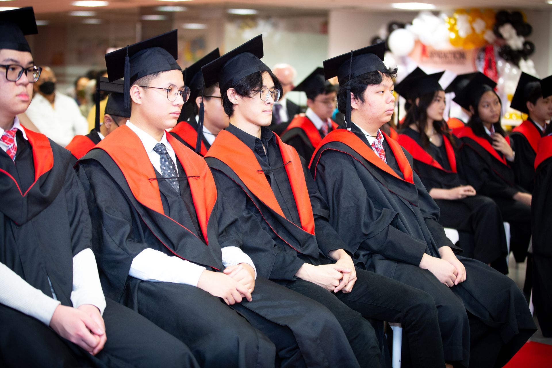 Students in graduation gowns and caps sit in a row during a ceremony with decorated balloons in the background.