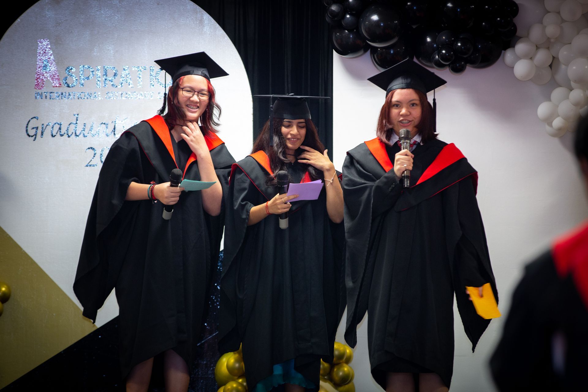 Three people in black graduation gowns and caps standing on a stage, with two holding microphones and one holding a paper.