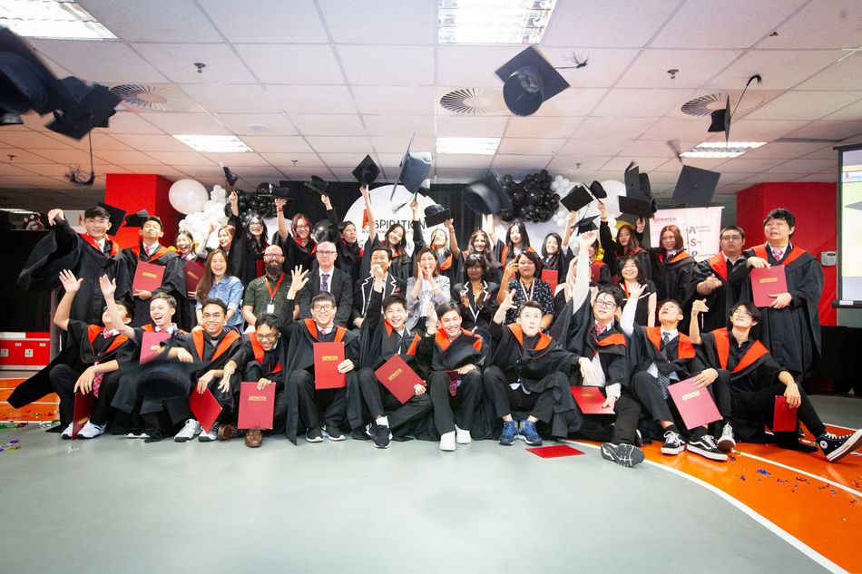 Graduates in academic regalia toss caps into the air at a ceremony, cheering and holding diplomas in an indoor hall.