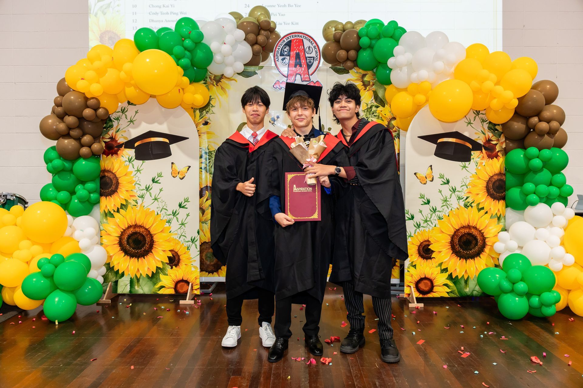 Three people in black graduation gowns and caps standing on a stage, with two holding microphones and one holding a paper.