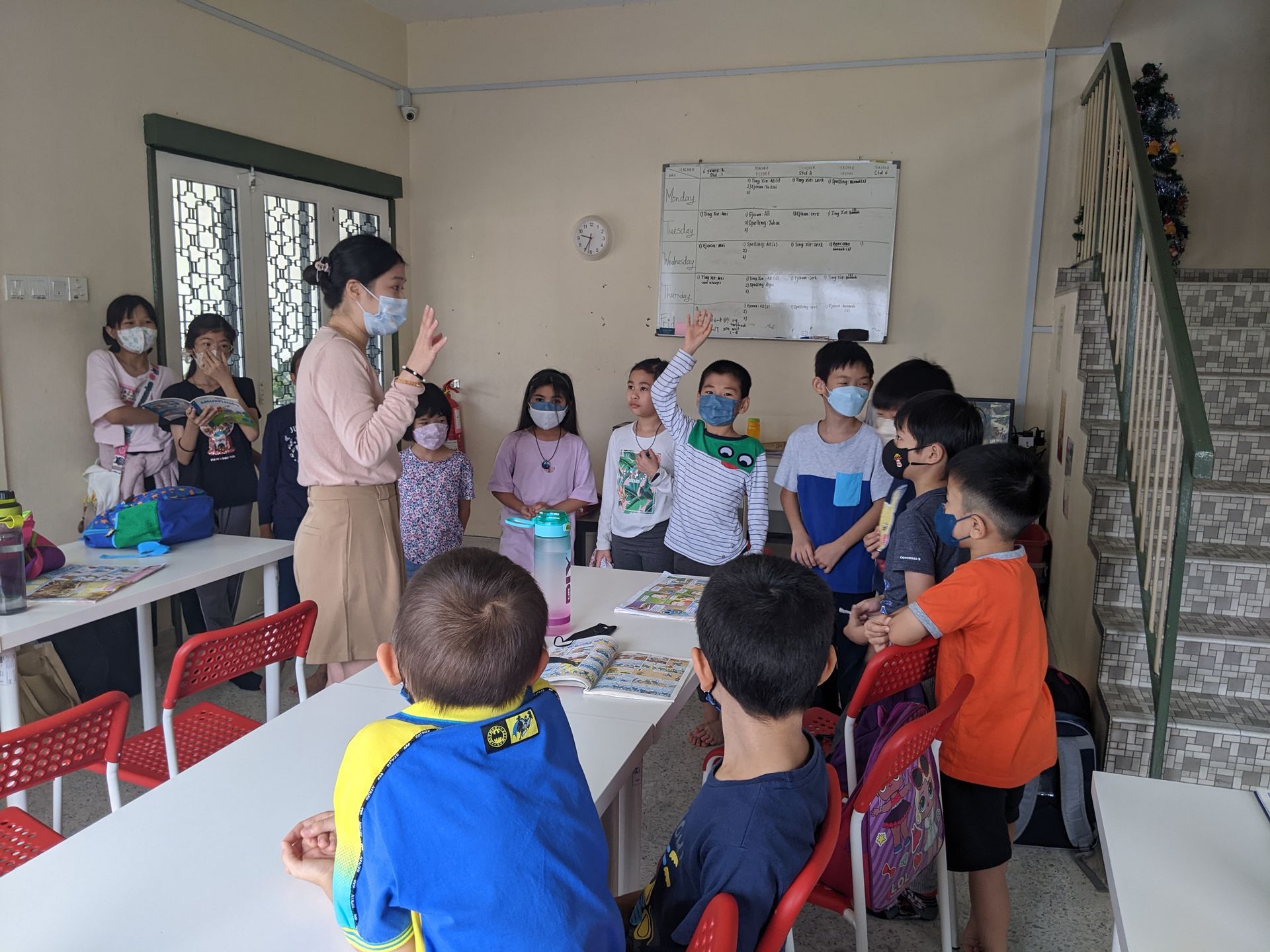 A teacher wearing a mask instructs a classroom of children, with some students raising their hands to participate.