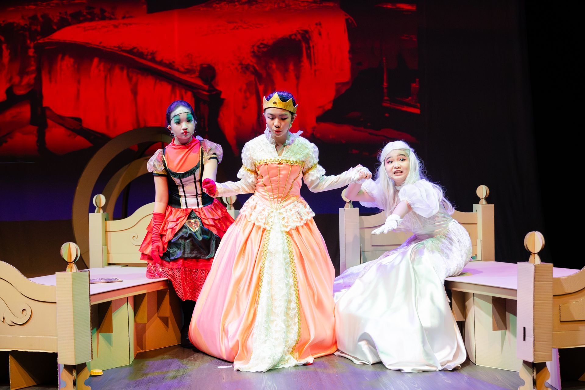 Three performers in costumes stand on stage between two wooden beds, holding hands in a theatrical scene.