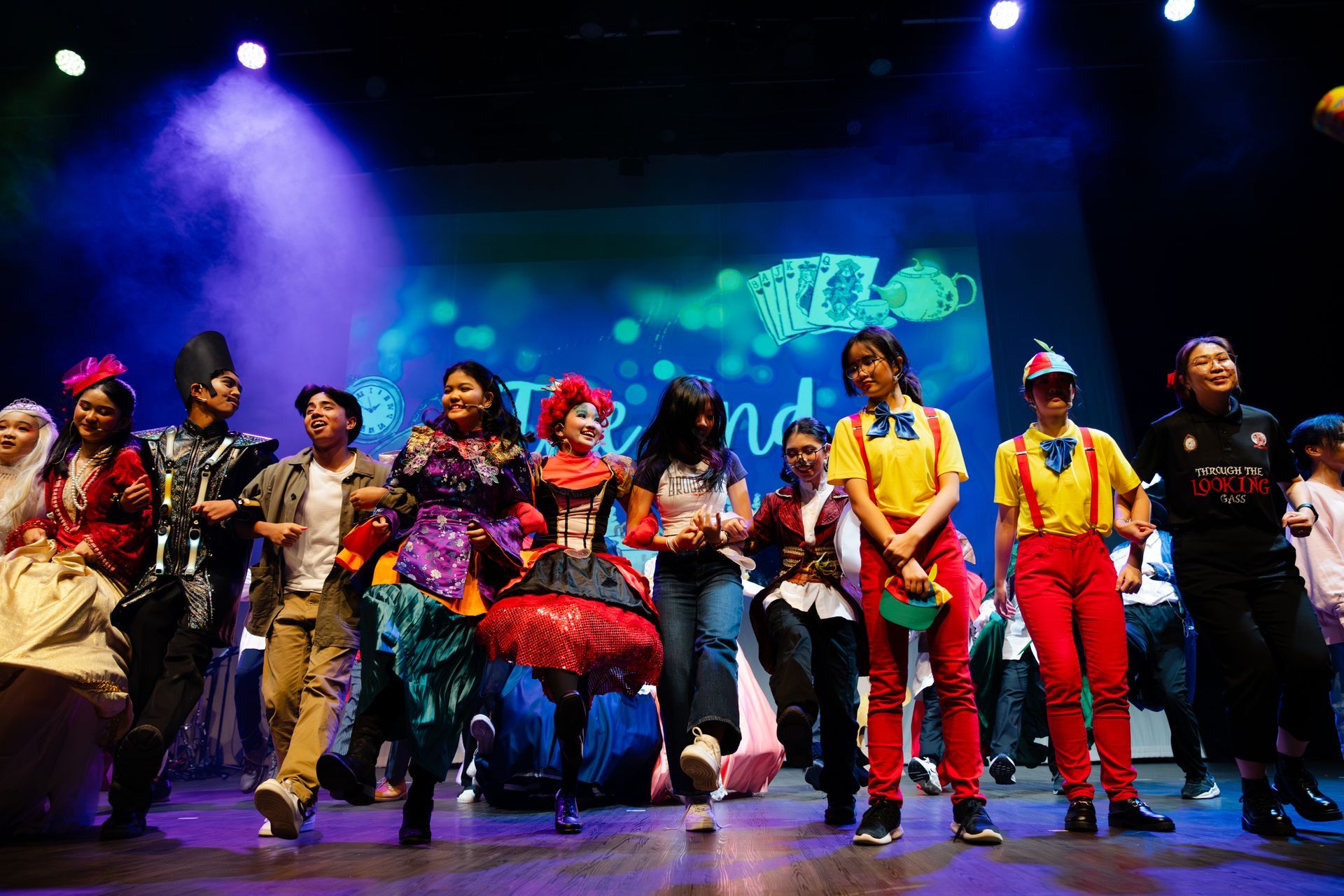 A group of performers in various colorful costumes dance and pose on a stage under bright, dramatic spotlighting.