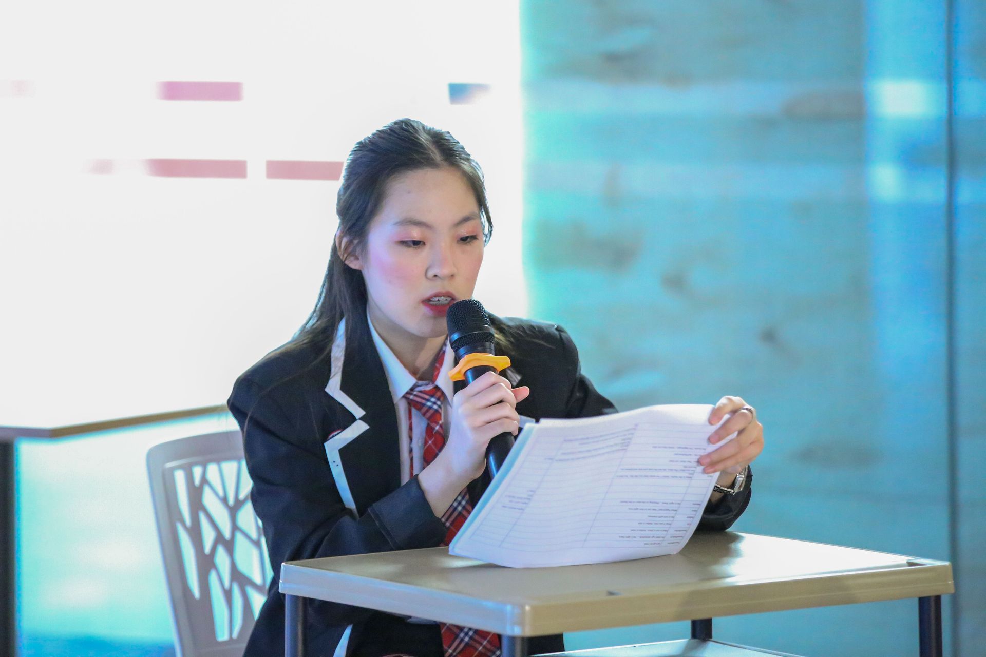 A person wearing a suit and tie speaks into a microphone while reading from a document at a desk.