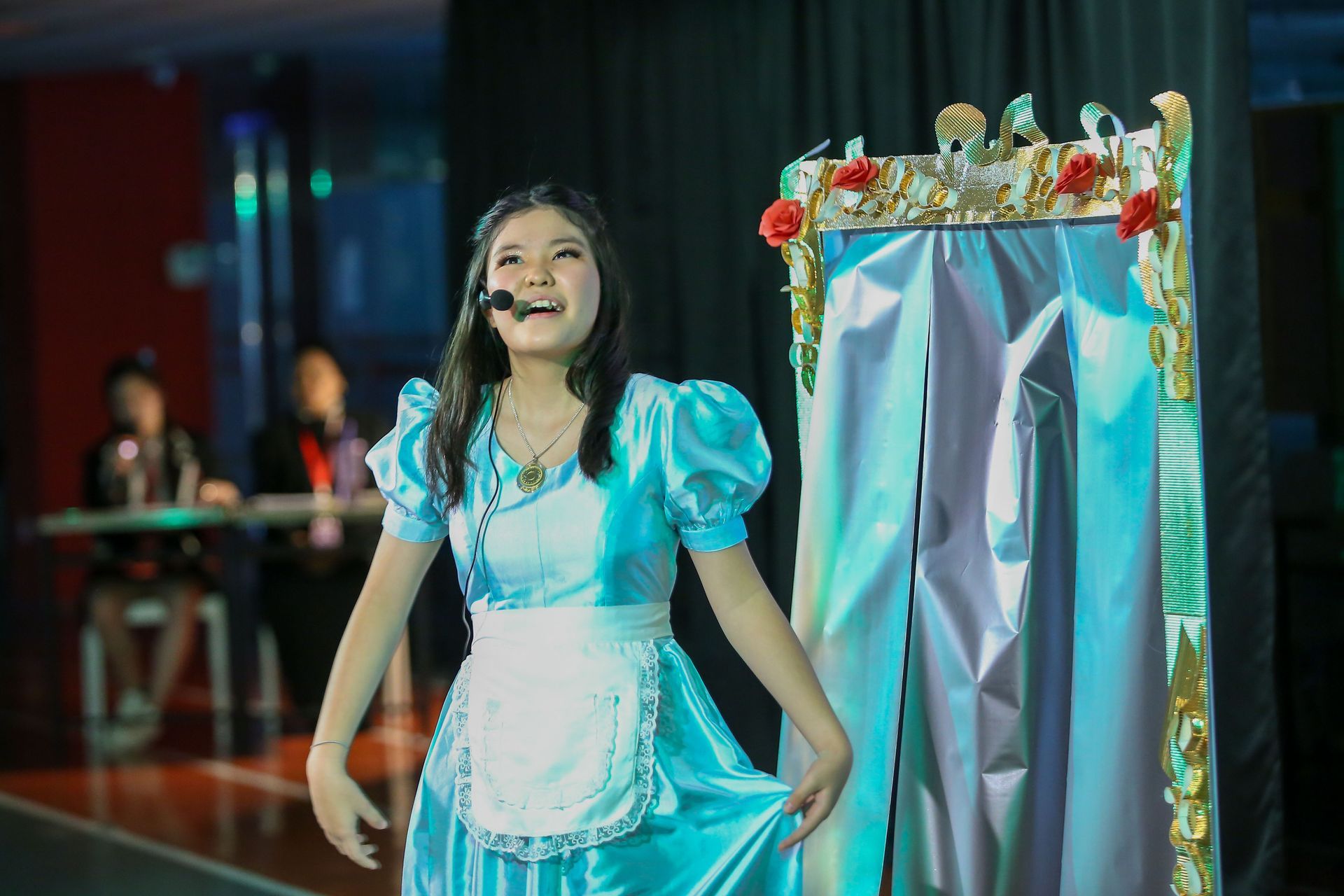 A performer in a blue dress with a white apron sings into a headset microphone next to a decorated, draped stage prop.