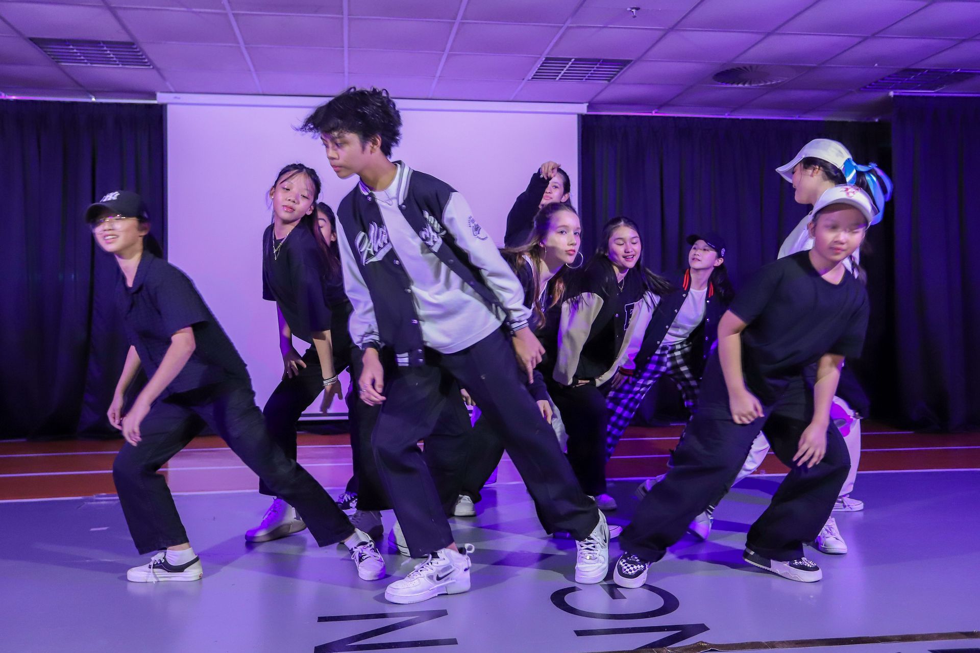A group of dancers in casual dark clothing and sneakers perform a synchronized routine on a stage with purple lighting.