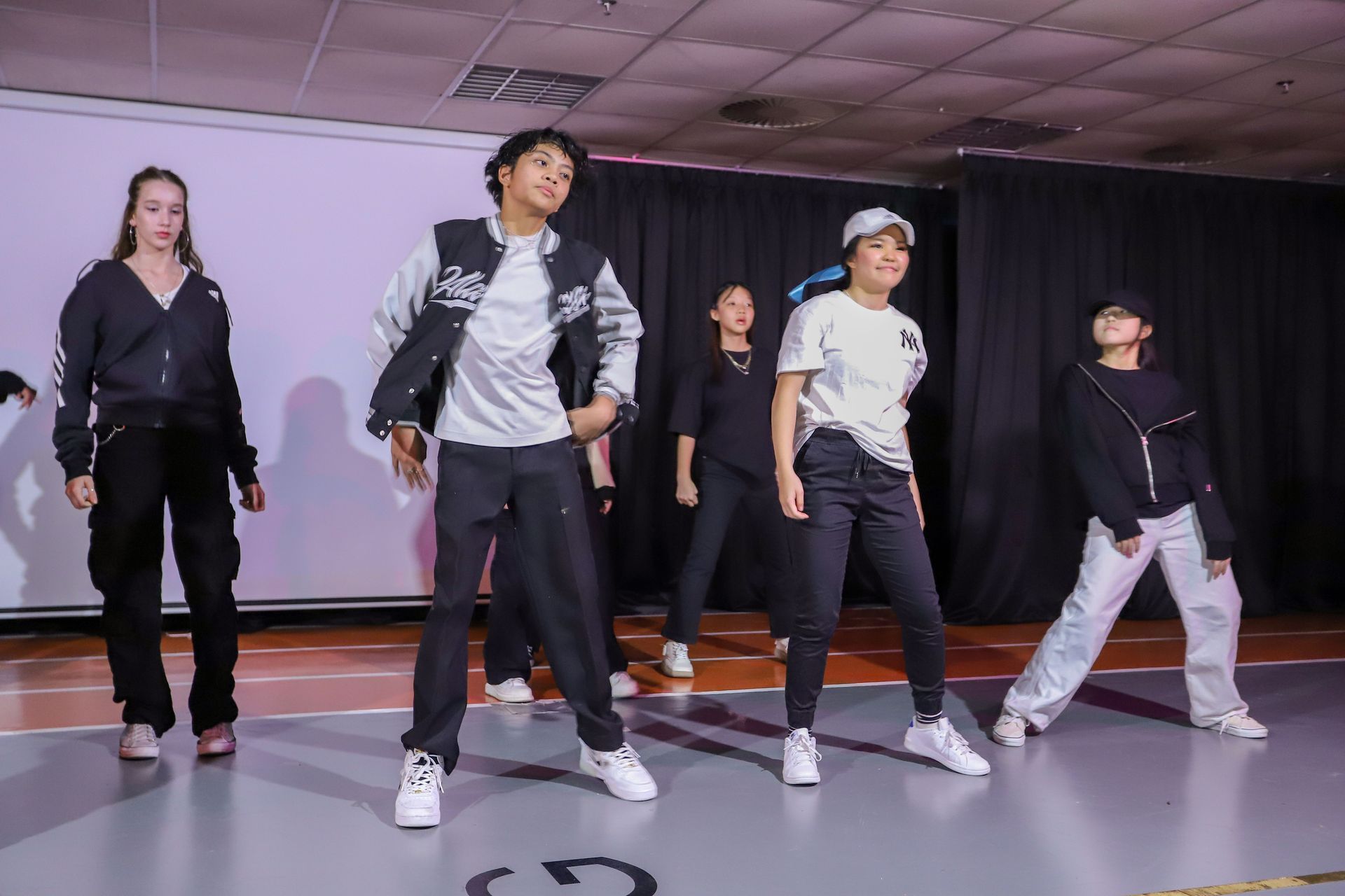 A group of five individuals practicing a synchronized dance routine in a studio with a white screen and black curtains.