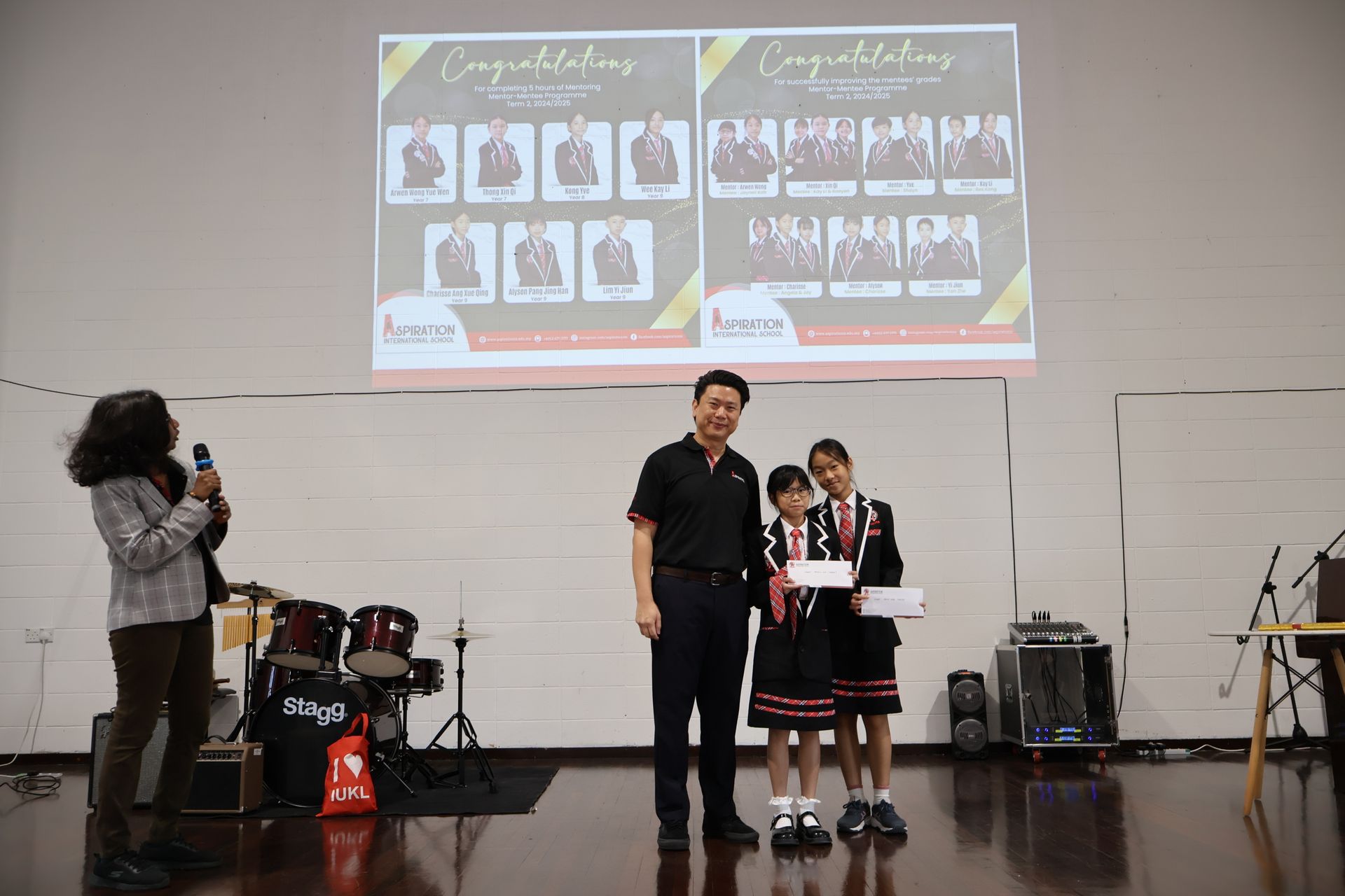 A person presents certificates to two students on a stage in front of a screen displaying award winners.