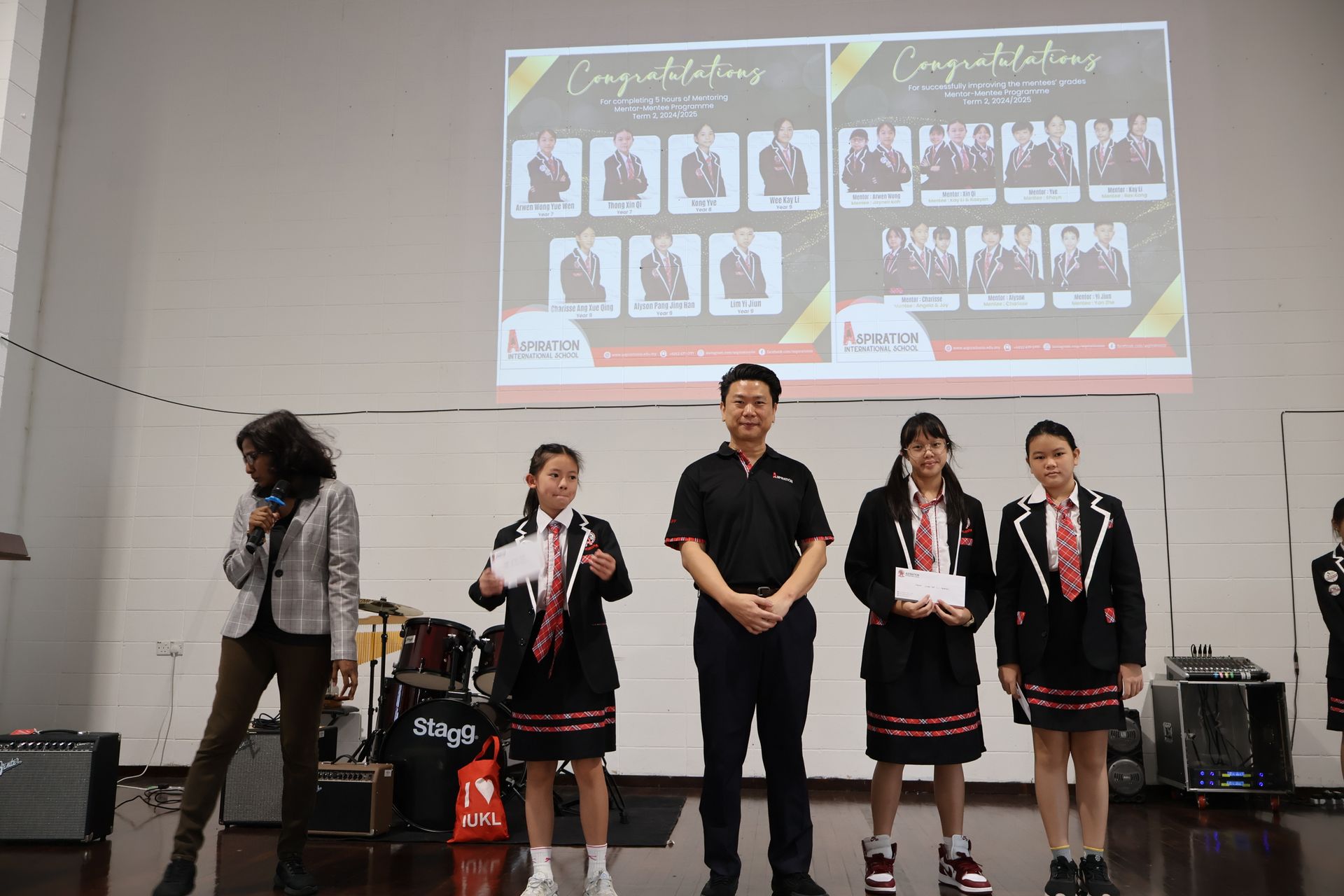 A group of students and an adult stand on a stage in front of a screen displaying congratulatory student photos.
