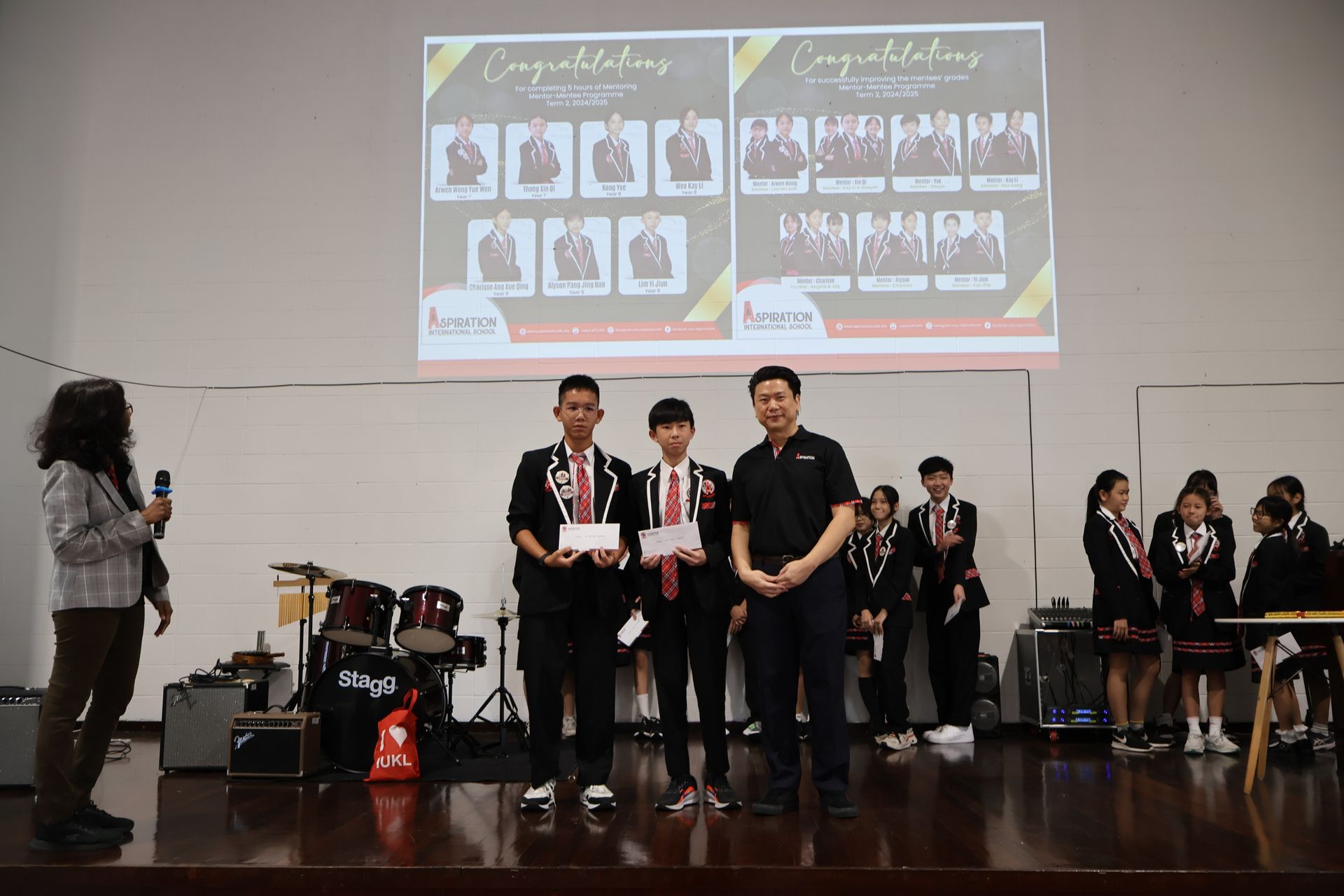 Students holding awards on a stage during an event, with a congratulatory presentation displayed on a screen behind them.