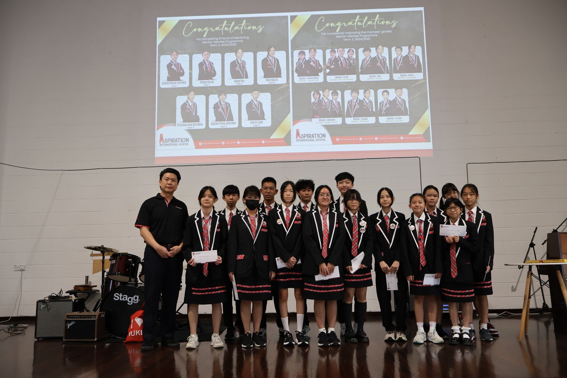 A group in school uniforms stands before a projector screen displaying student award winners during an indoor ceremony.