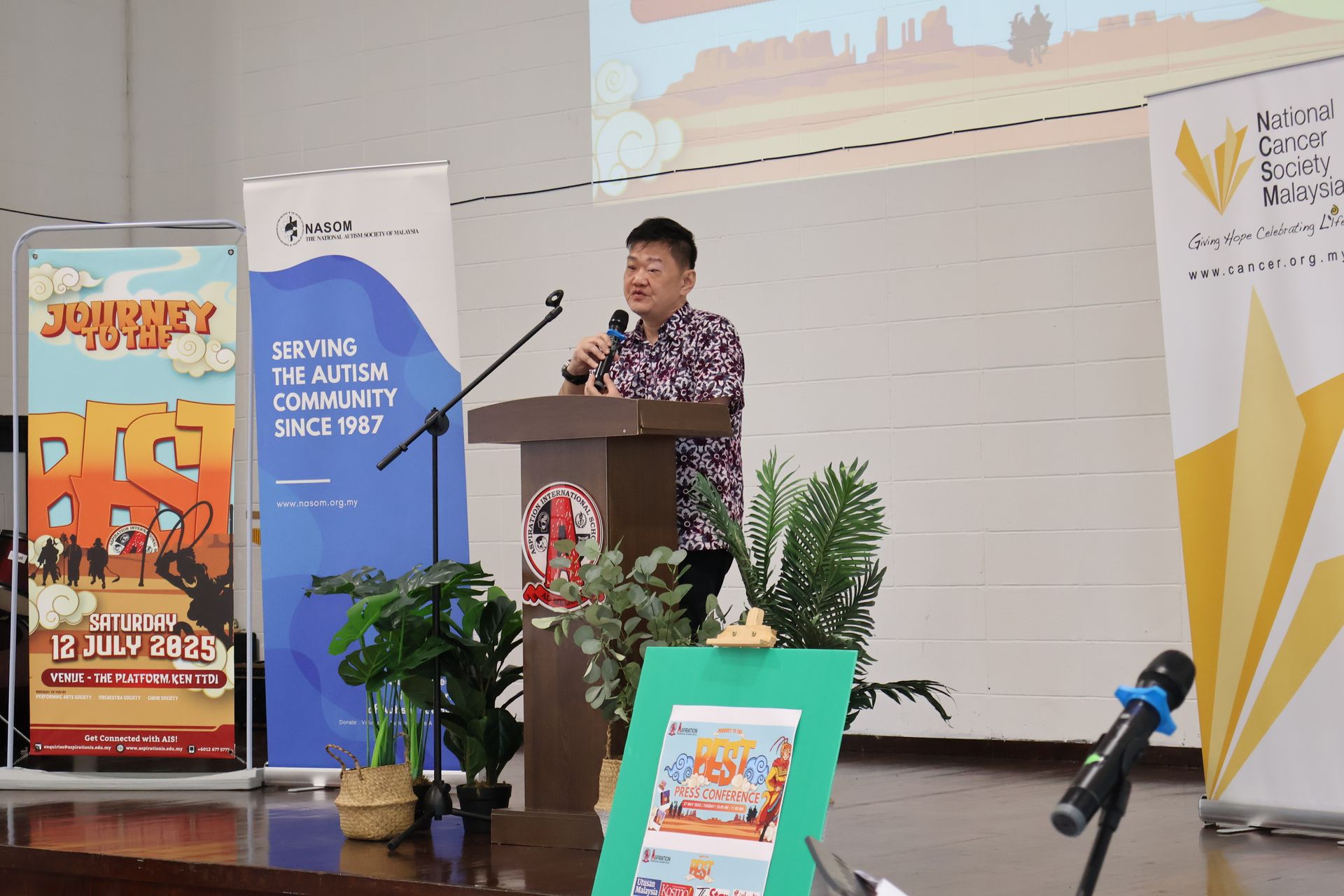 A person standing at a podium speaking, with event banners and plants on a stage in front of a projection screen.