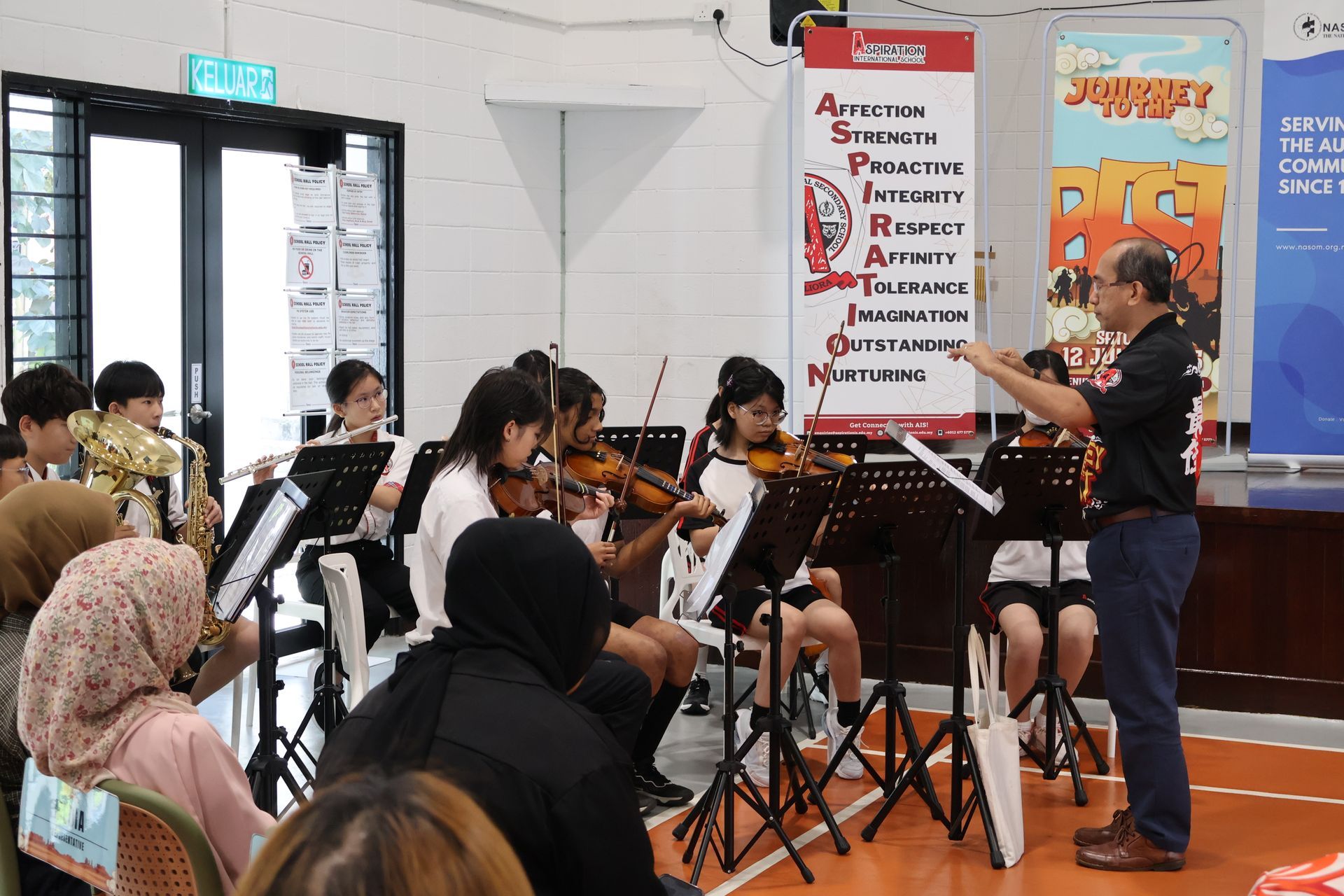 A conductor leads an orchestra of students playing string and wind instruments during a rehearsal in a school hall.