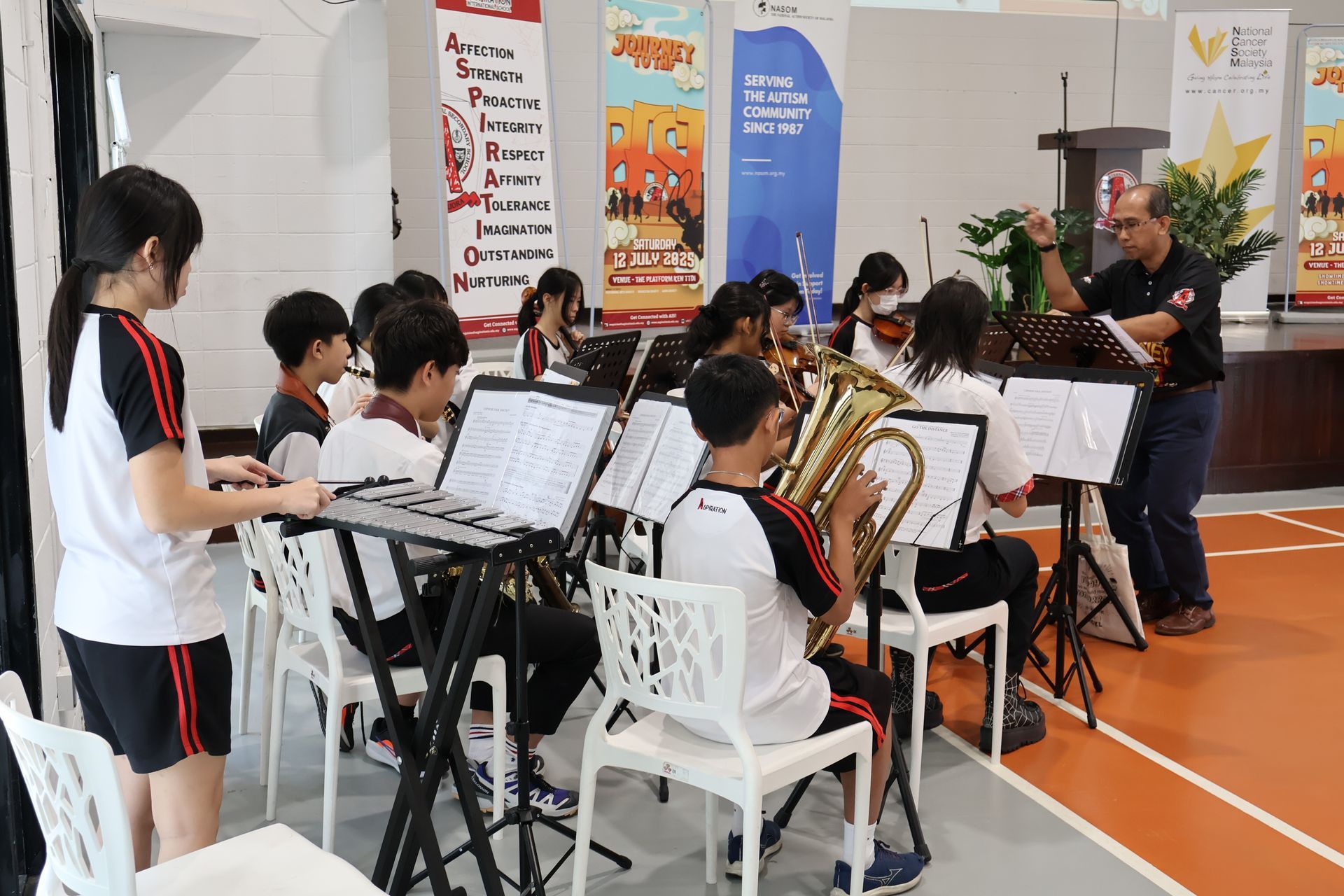 Students in uniforms play musical instruments in an orchestra rehearsal, led by a conductor in a gym.
