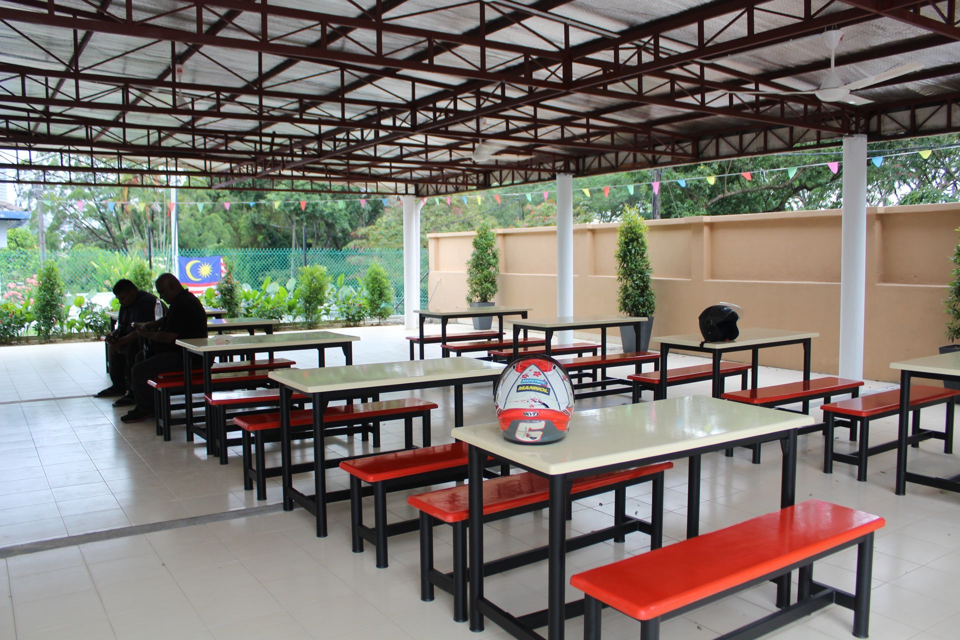 An open-air dining area with several tables and red benches, featuring a helmet on one table and a person seated nearby.