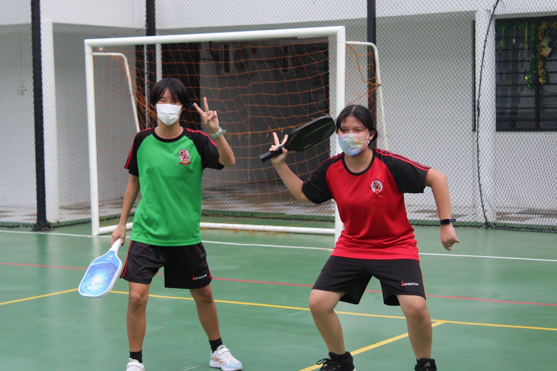 Two people in sports attire holding pickleball paddles pose on a court in front of a goal net while wearing face masks.