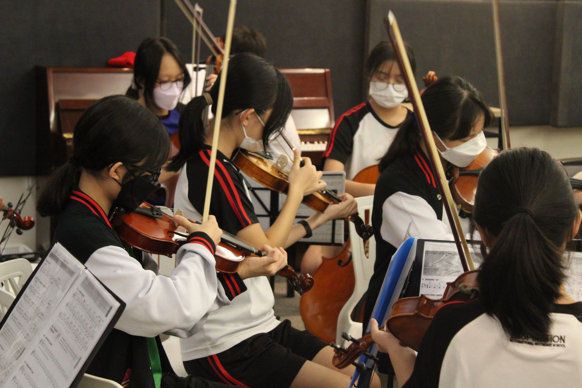 Students in matching athletic uniforms play violins during an orchestral rehearsal in a practice room.