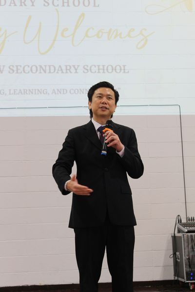 A person in a black suit holds a microphone, speaking to an audience in front of a school welcome sign.
