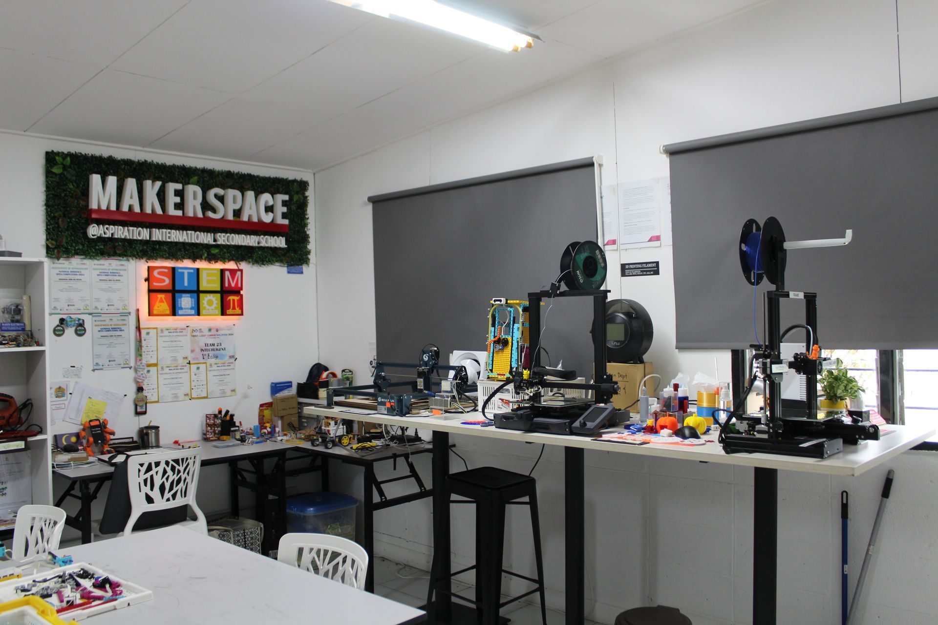 A makerspace room featuring 3D printers on tables, chairs, a white table in the foreground, and a large wall sign.