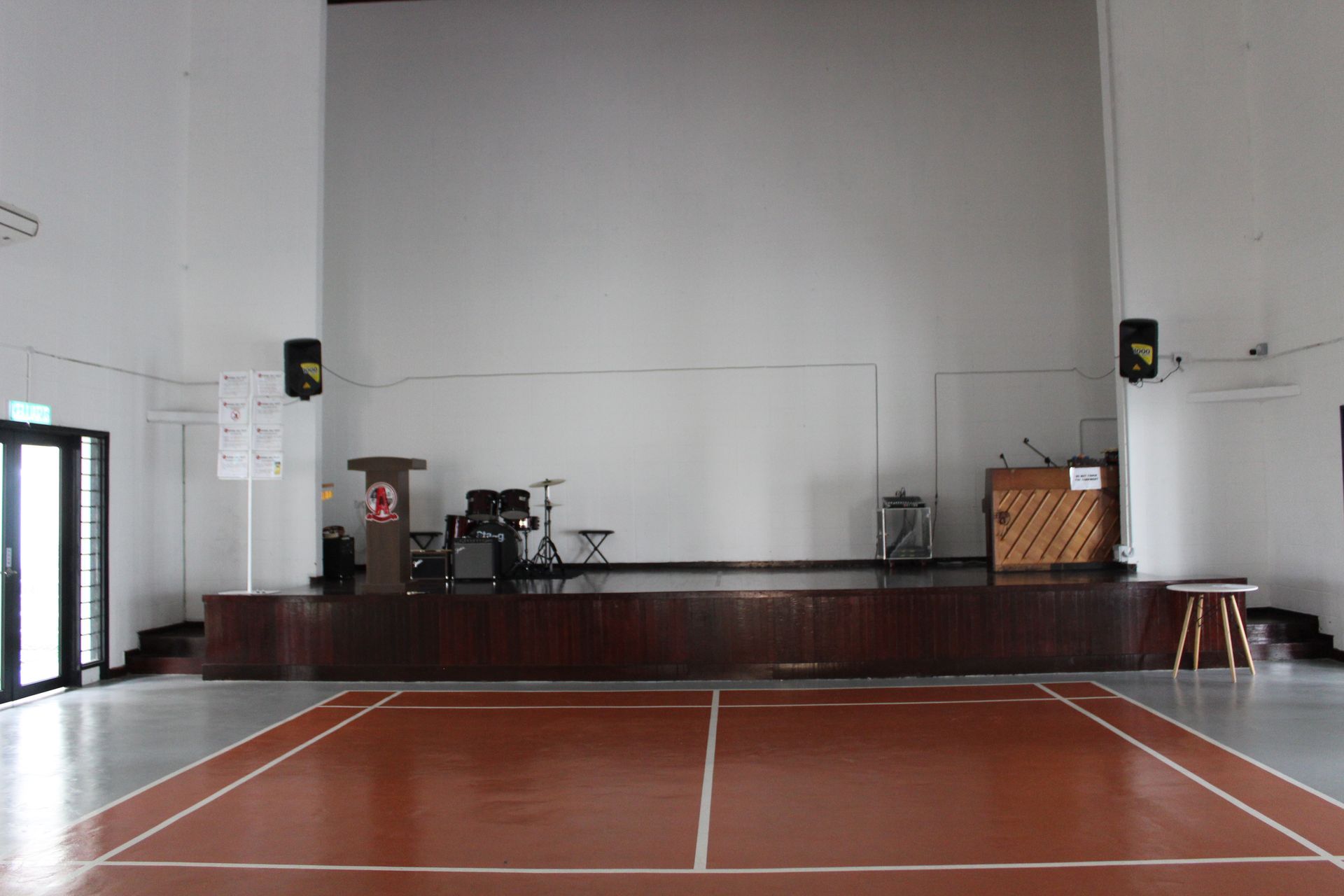 A school hall featuring a wooden stage with audio equipment, a podium, and a brown badminton court on the floor.