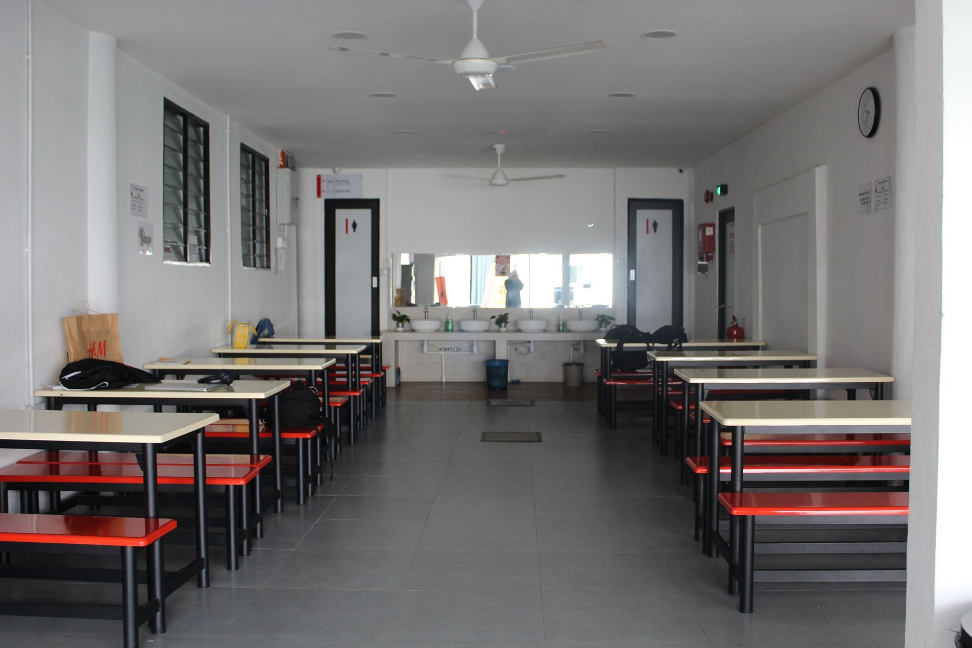 A cafeteria with rows of tables, red benches, white walls, and a wash station at the far end of the room.