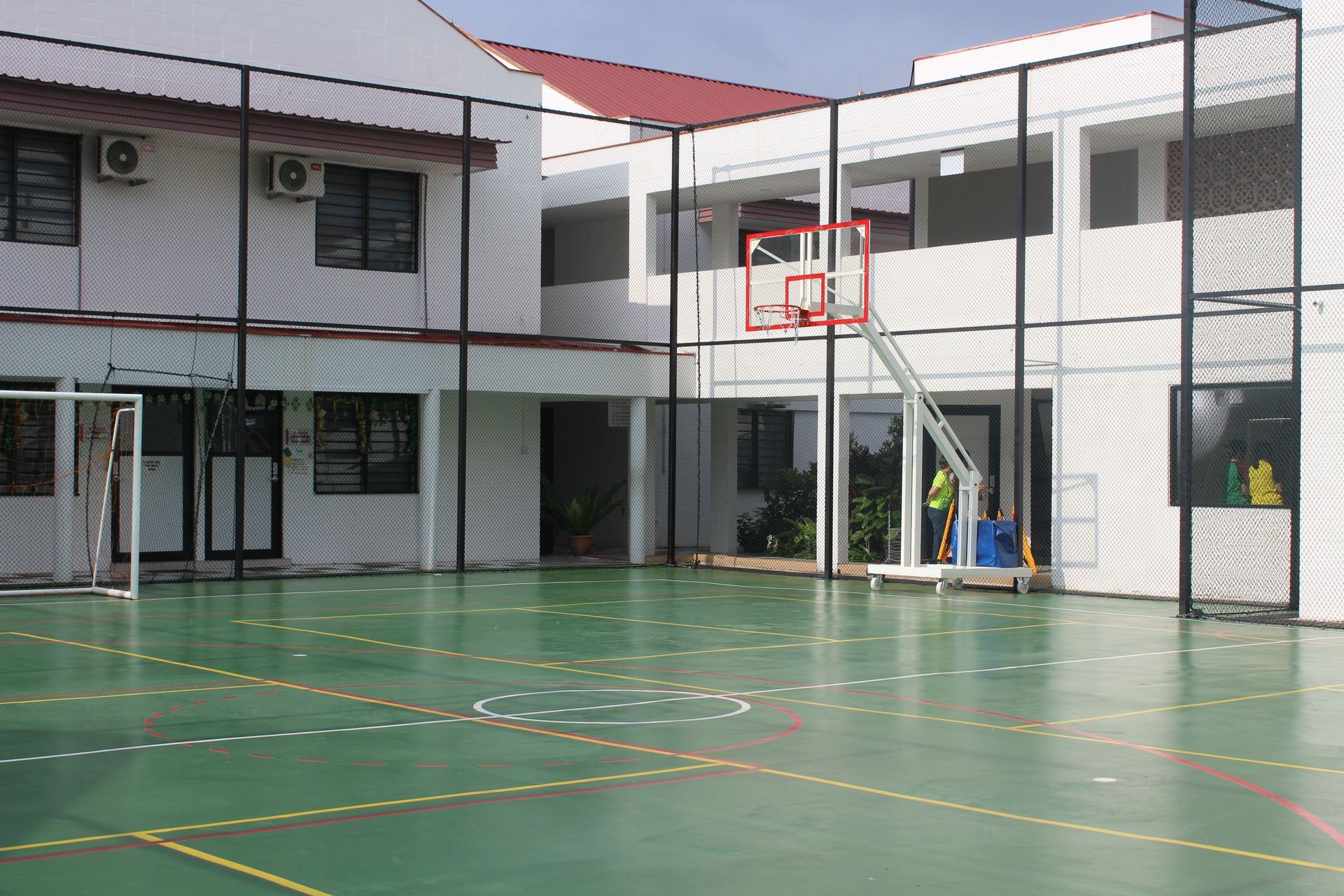 An outdoor sports court with a basketball hoop, green surface, and white boundary lines, set between school buildings.