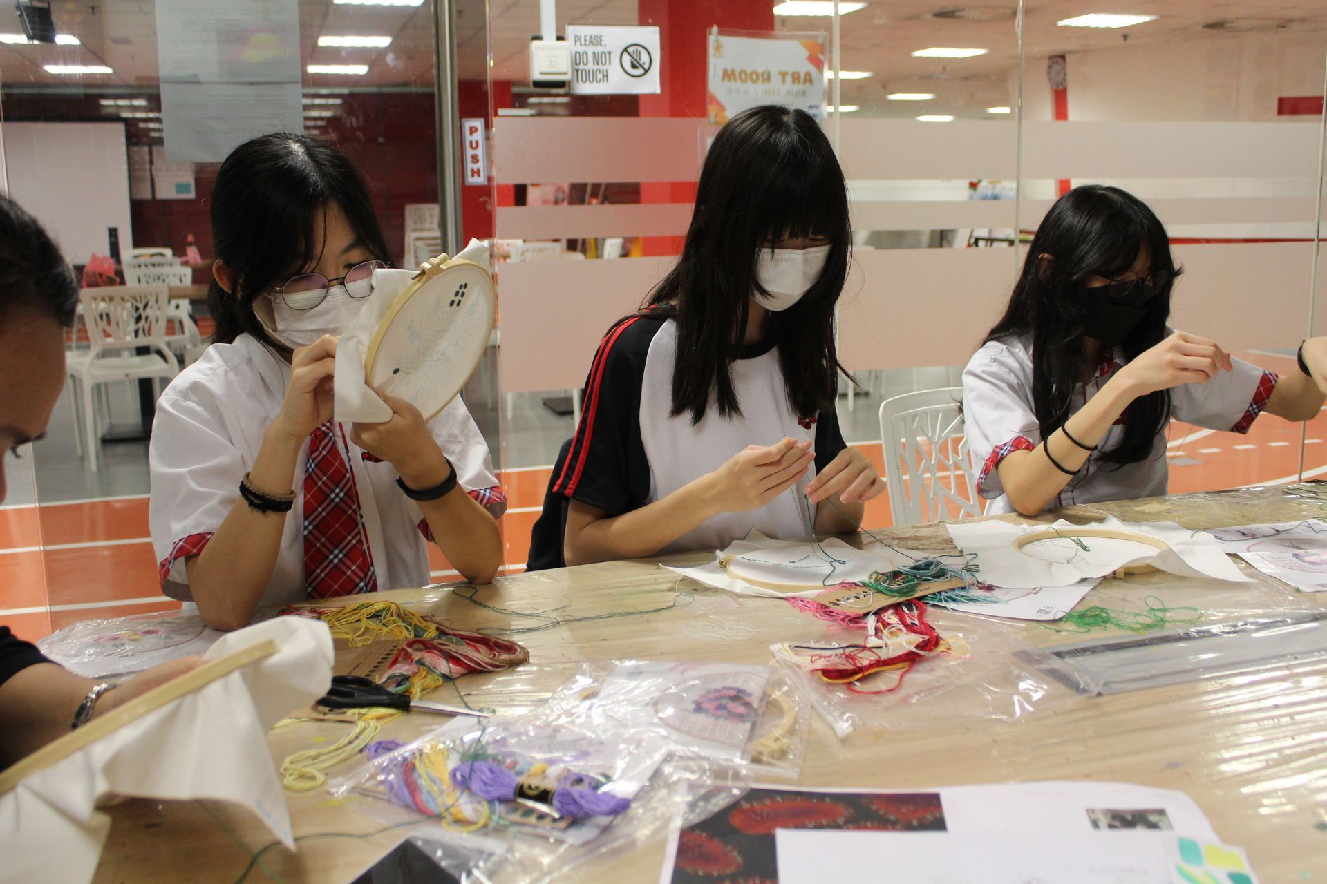 Three students wearing face masks sit at a table embroidering fabric with hoops in a brightly lit indoor workspace.
