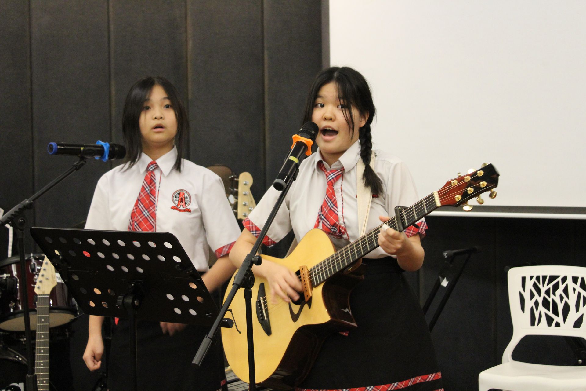 Two students in white school uniforms with plaid ties perform on stage, one playing acoustic guitar and both singing.