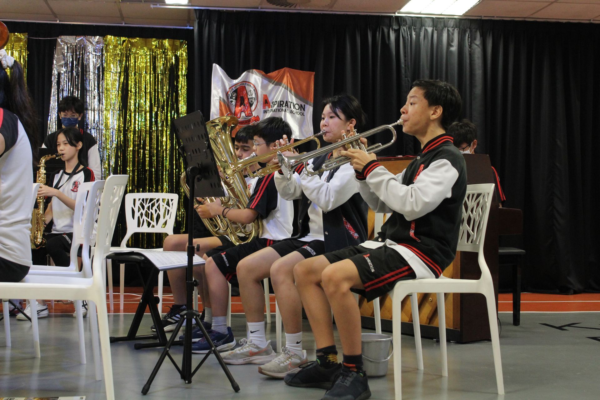 A music ensemble sits on white chairs, performing together while playing brass and woodwind instruments indoors.