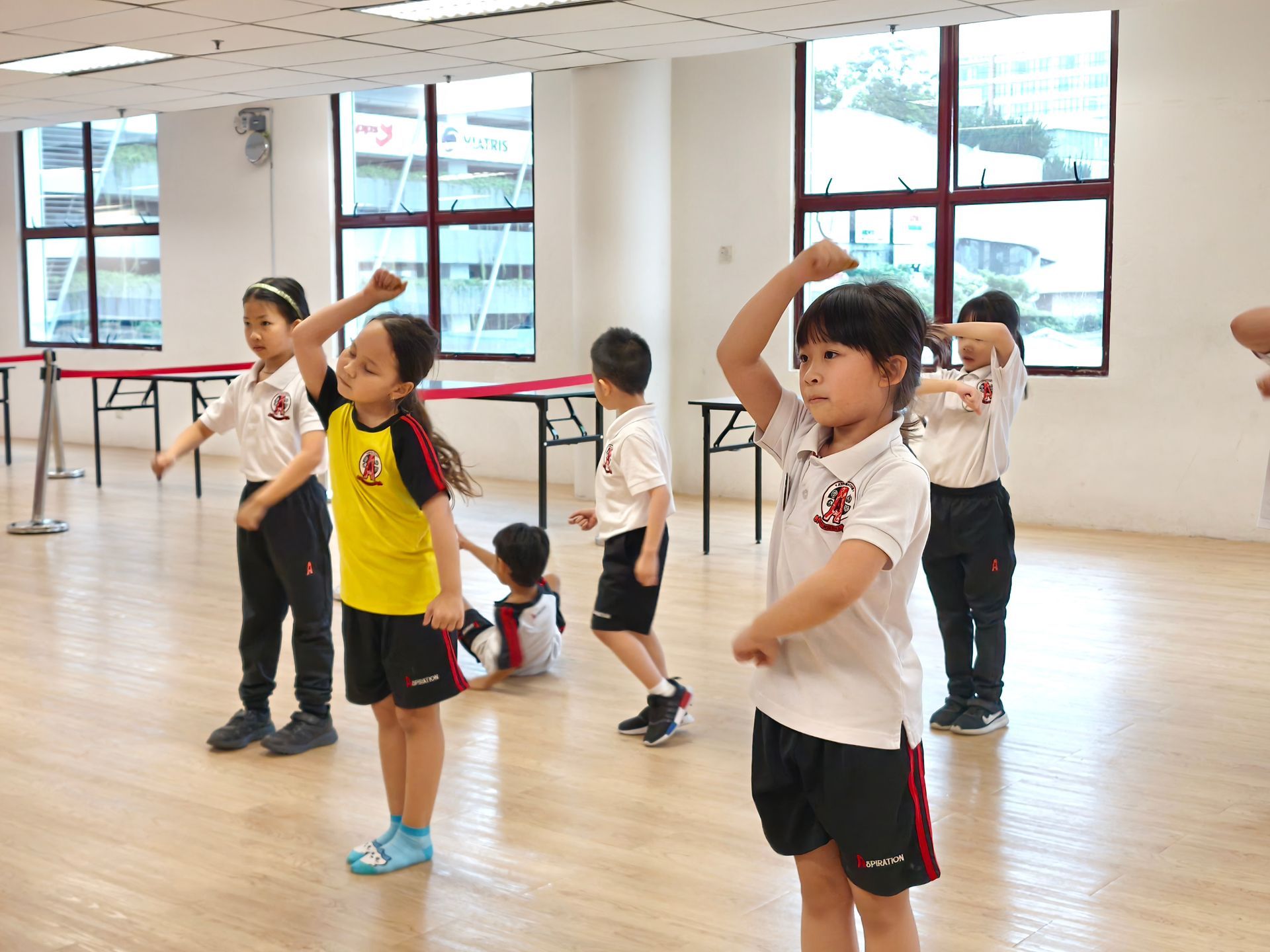 A group of children in uniform practicing dance moves in a bright studio with large windows and wooden floors.